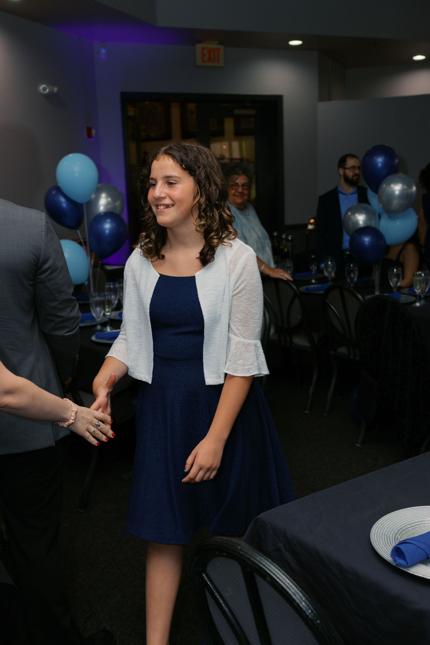 A smiling girl in a navy dress and white cardigan at a blue and silver balloon-decorated party venue.