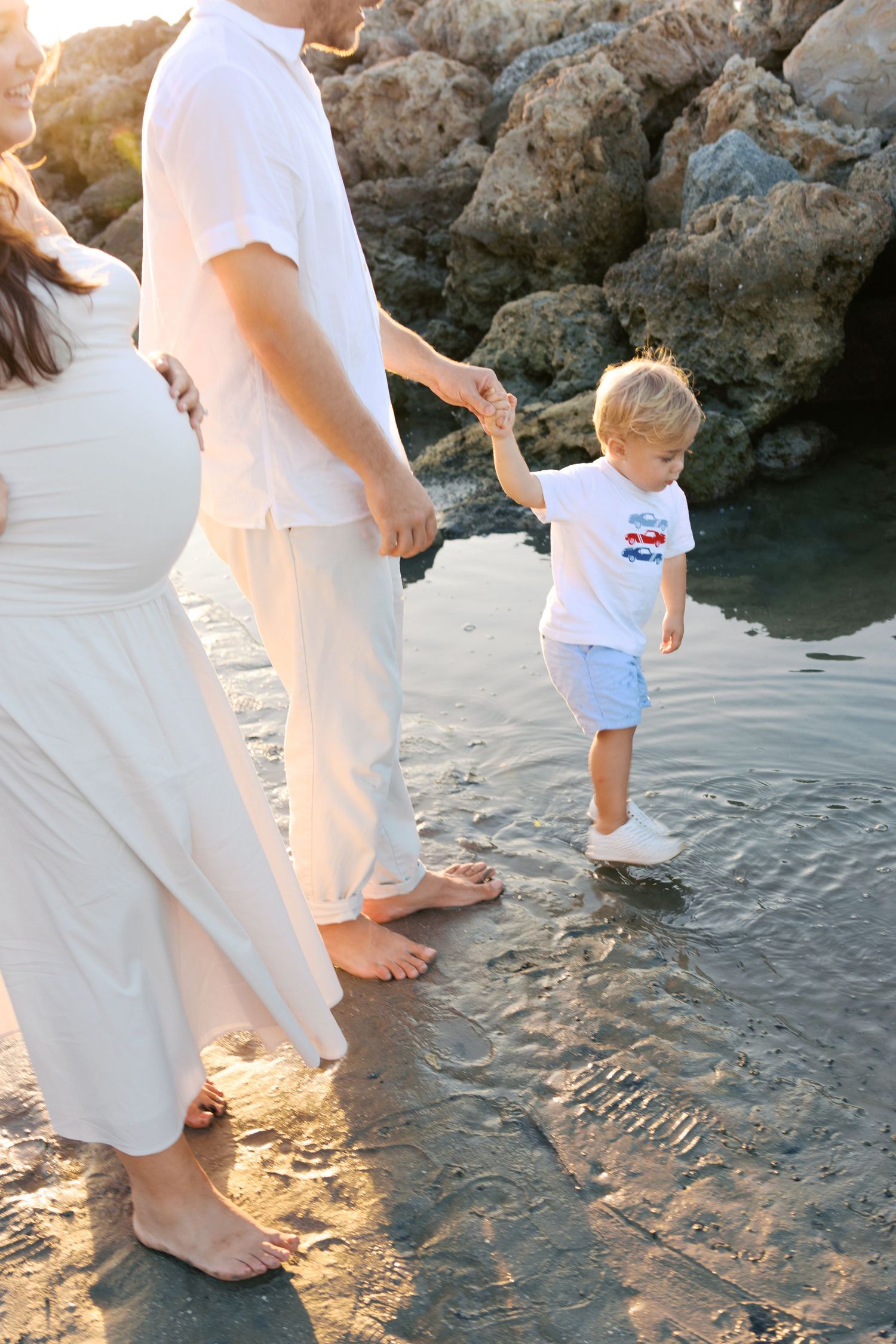 Pregnant woman and man hold toddler boy's hand while walking barefoot on a rocky beach at sunset.