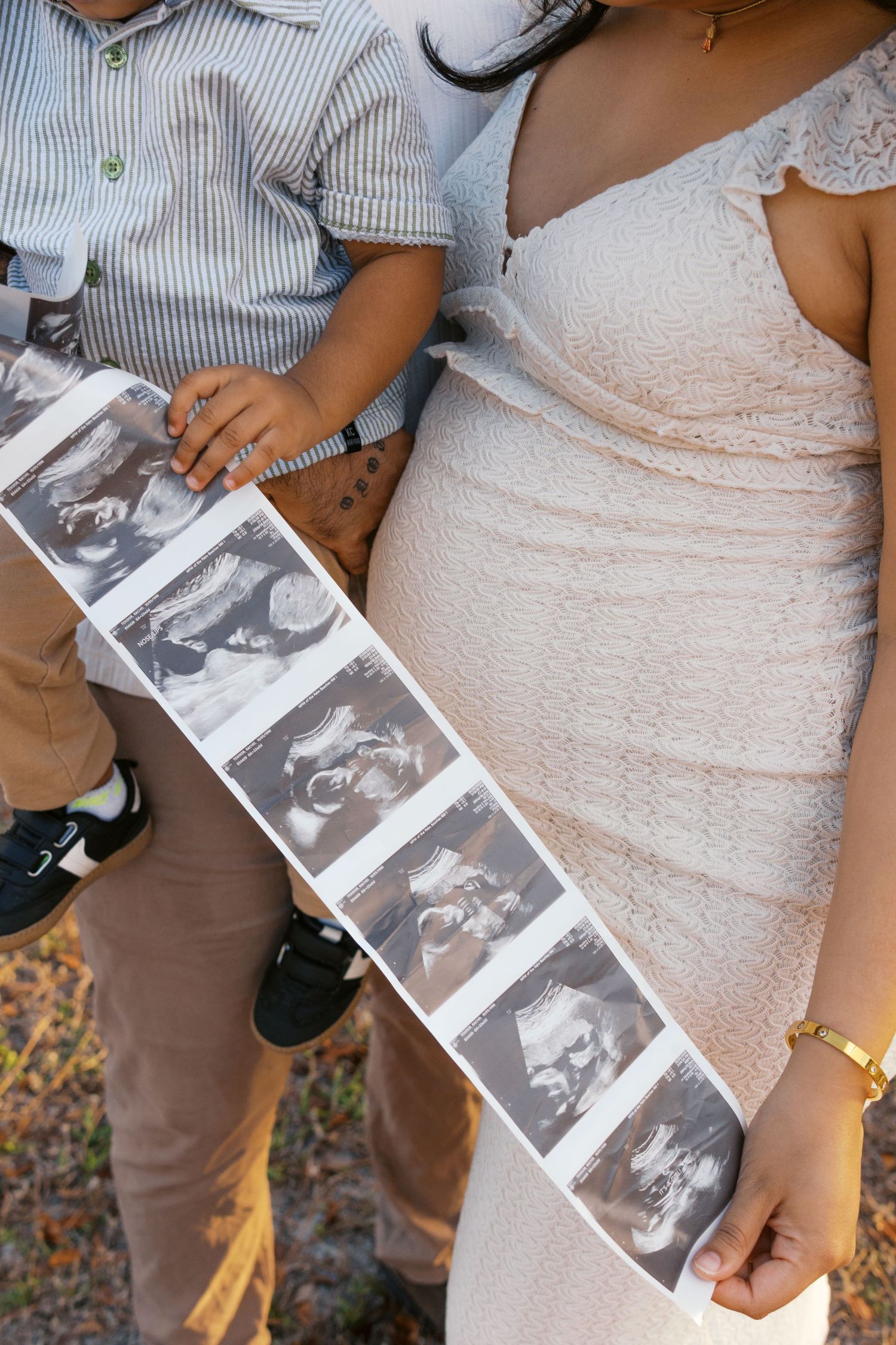 Pregnant woman in pink lace dress holds ultrasound strip while young child sits beside her outdoors.