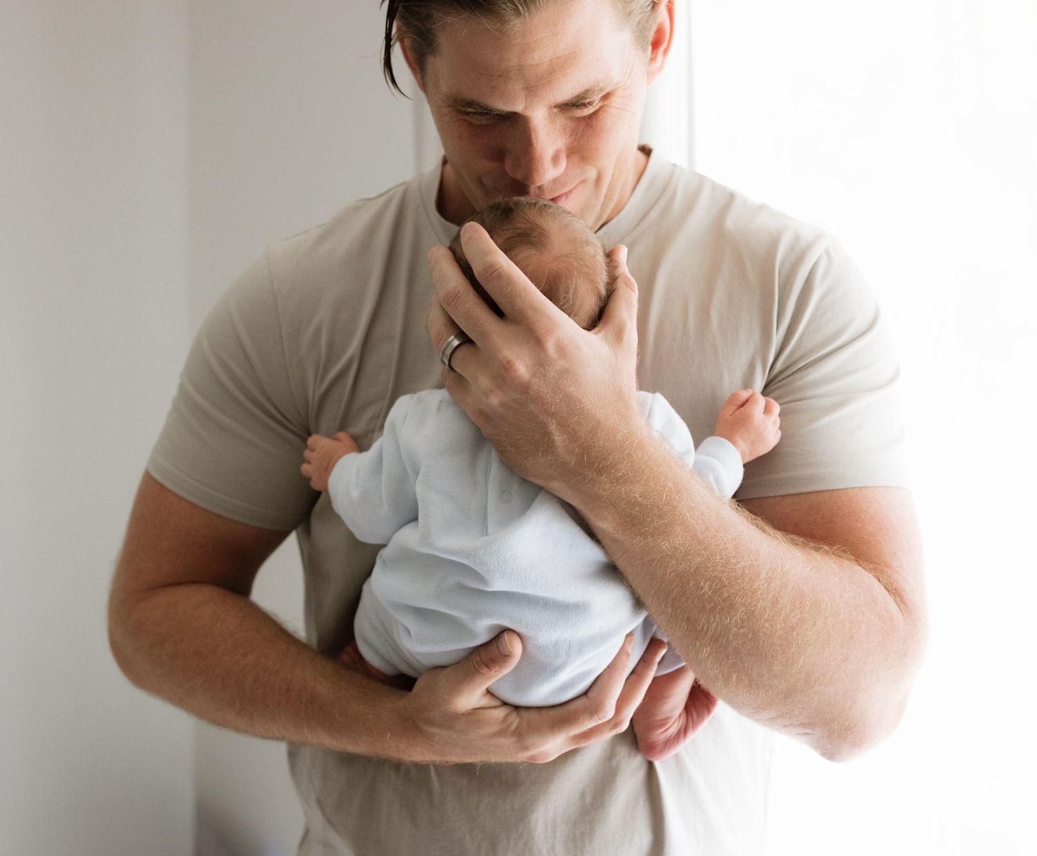 Parent holding sleeping baby in white onesie against neutral background.