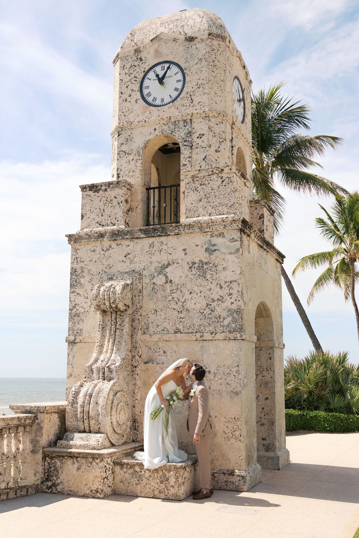 Bride in white gown poses at historic Palm Beach clock tower with ocean view and palm trees.