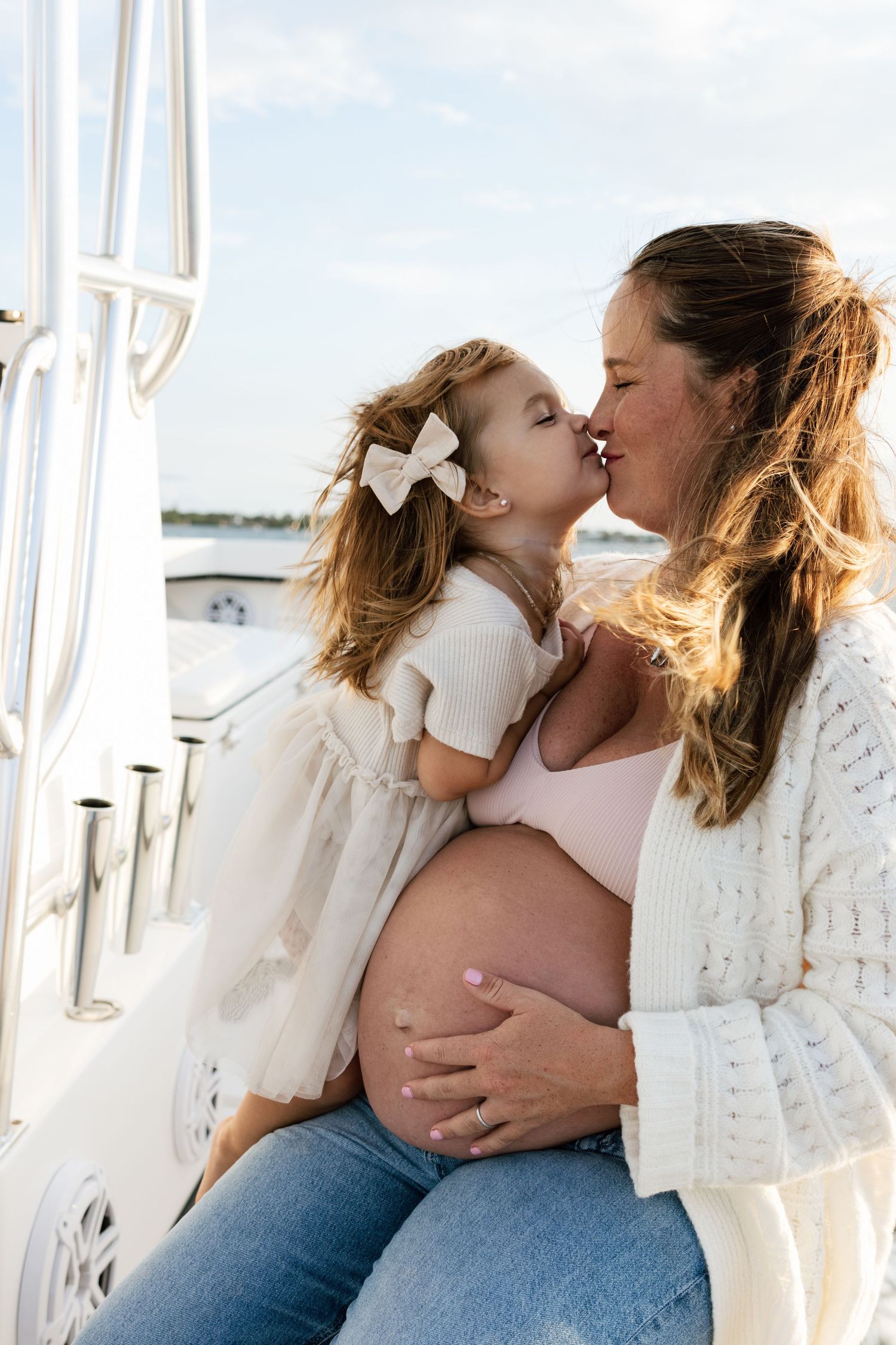 Pregnant woman sitting on a boat shares a tender kiss with her young daughter wearing a white bow and dress.