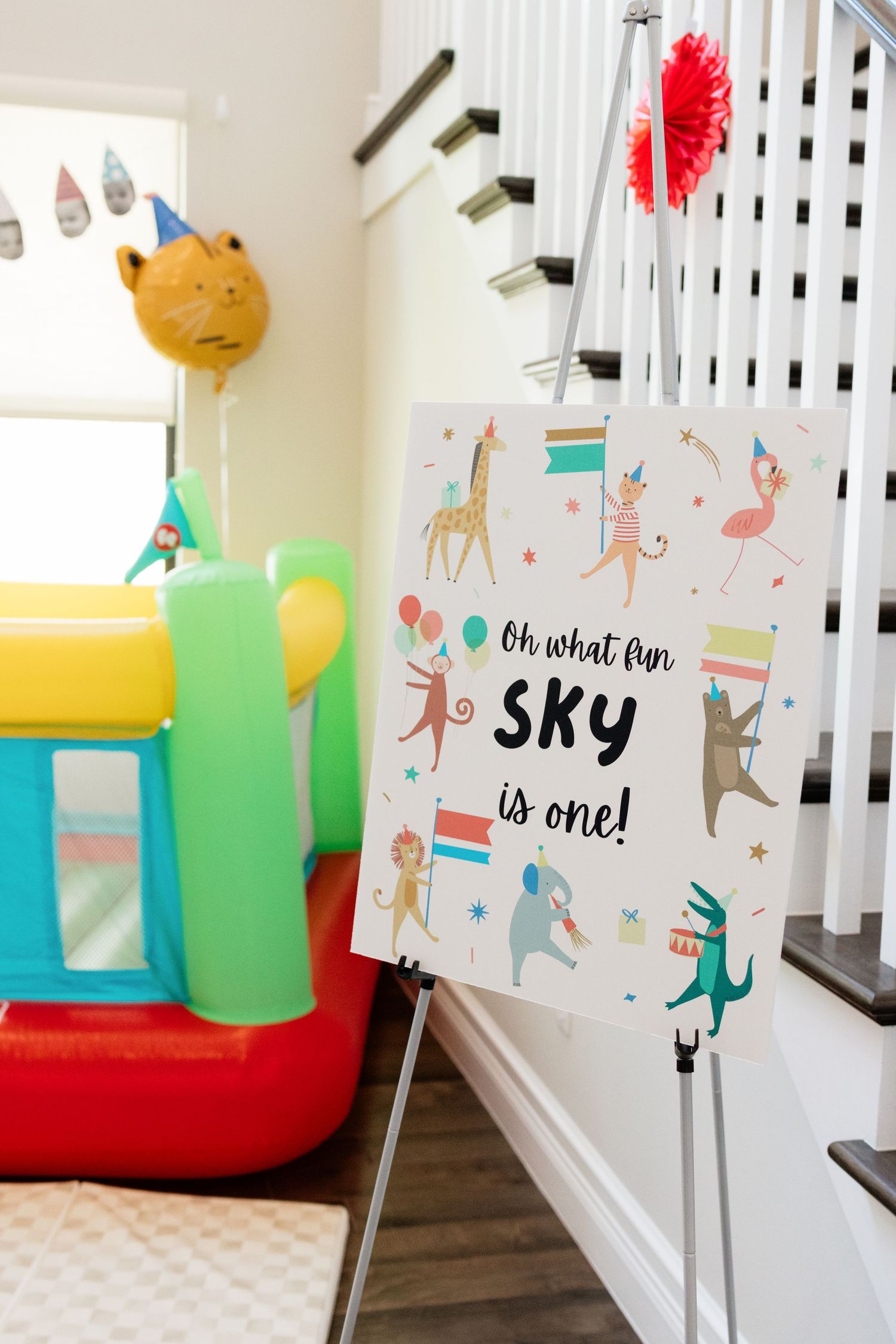 Colorful first birthday party setup with an easel sign reading Oh What Fun Sky is One, featuring festive animals and a bounce house.