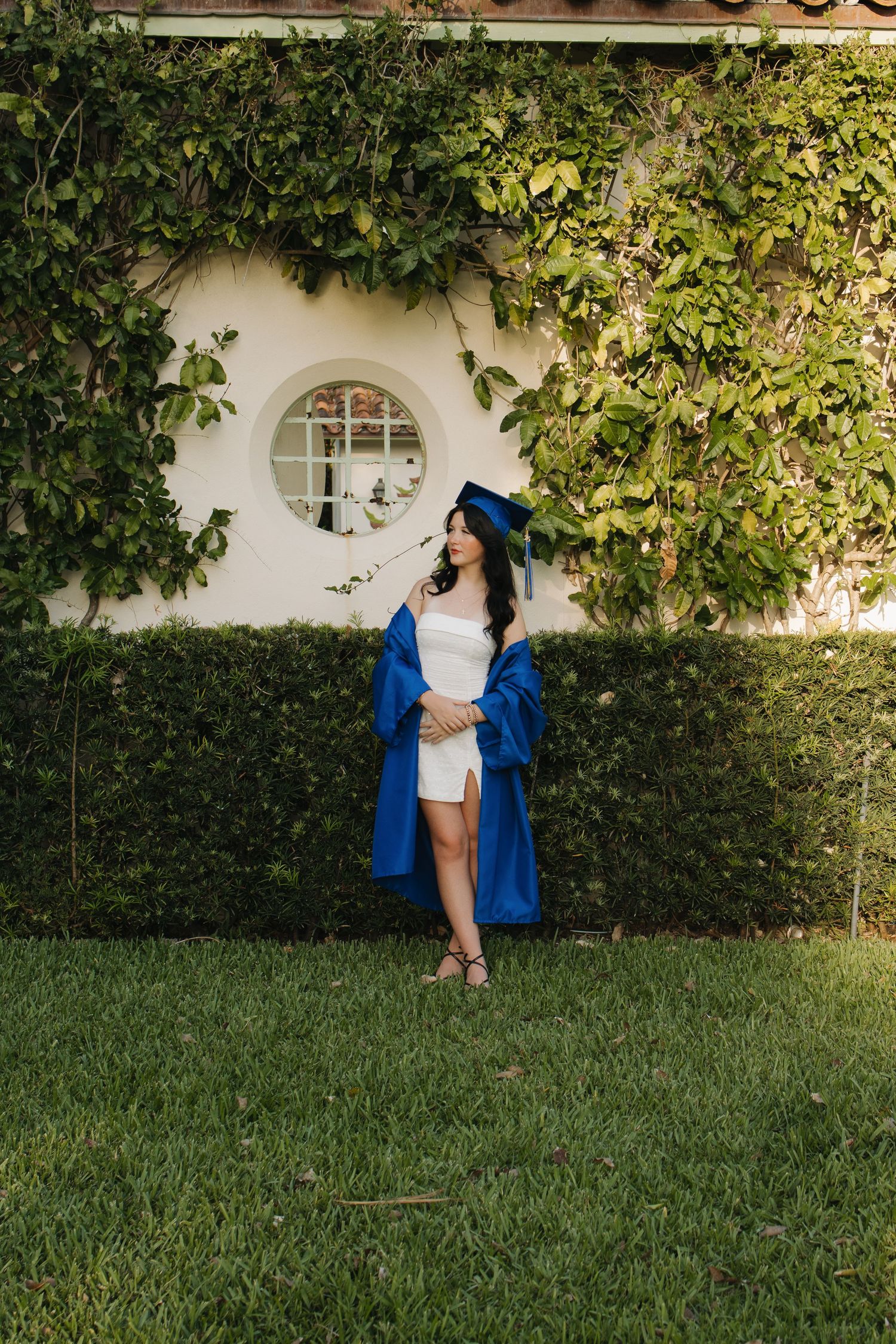Graduate in blue cap and gown poses against ivy-covered wall with round window.