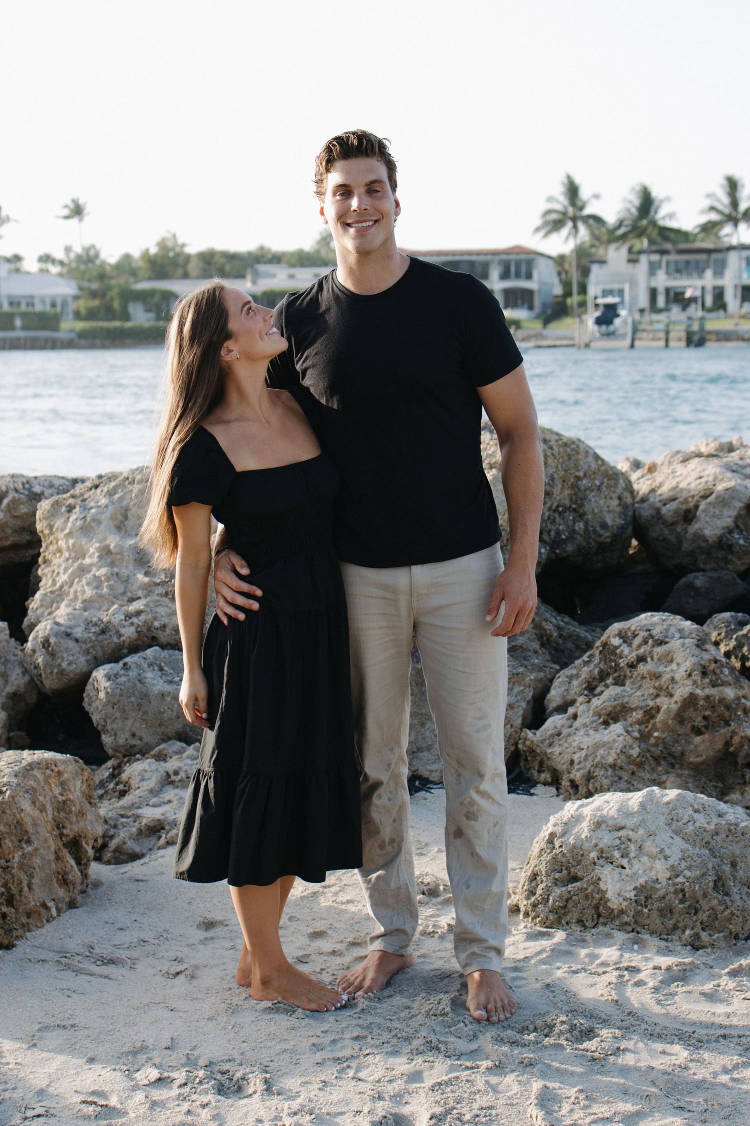 A couple poses together on a beach with rocks and waterfront homes in the background.