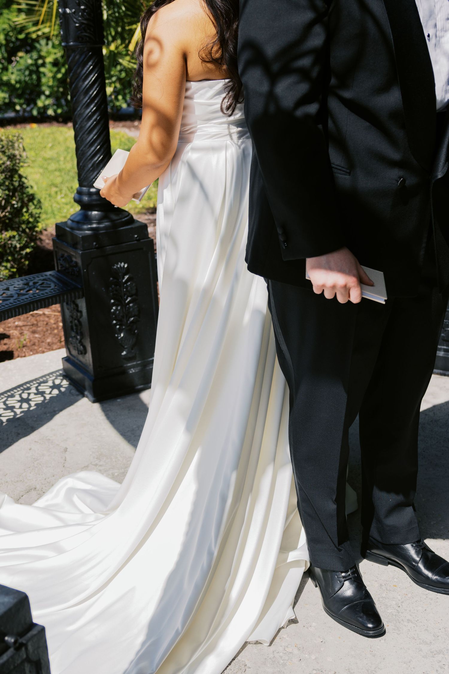 Bride in elegant white satin gown with train stands beside groom in black tuxedo outdoors at wedding ceremony.