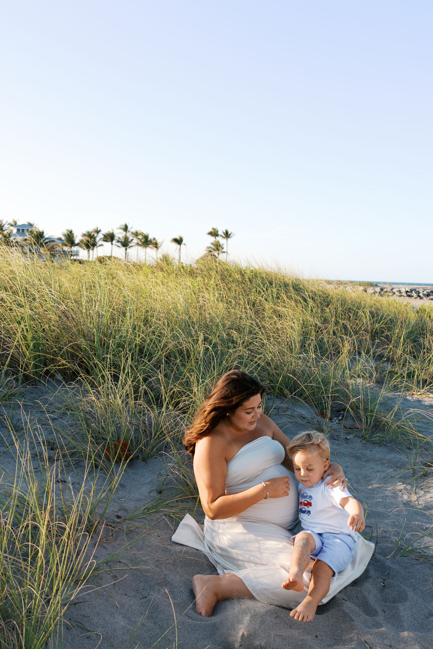 Pregnant woman in white dress sitting on beach sand dunes with young child, palm trees in background.