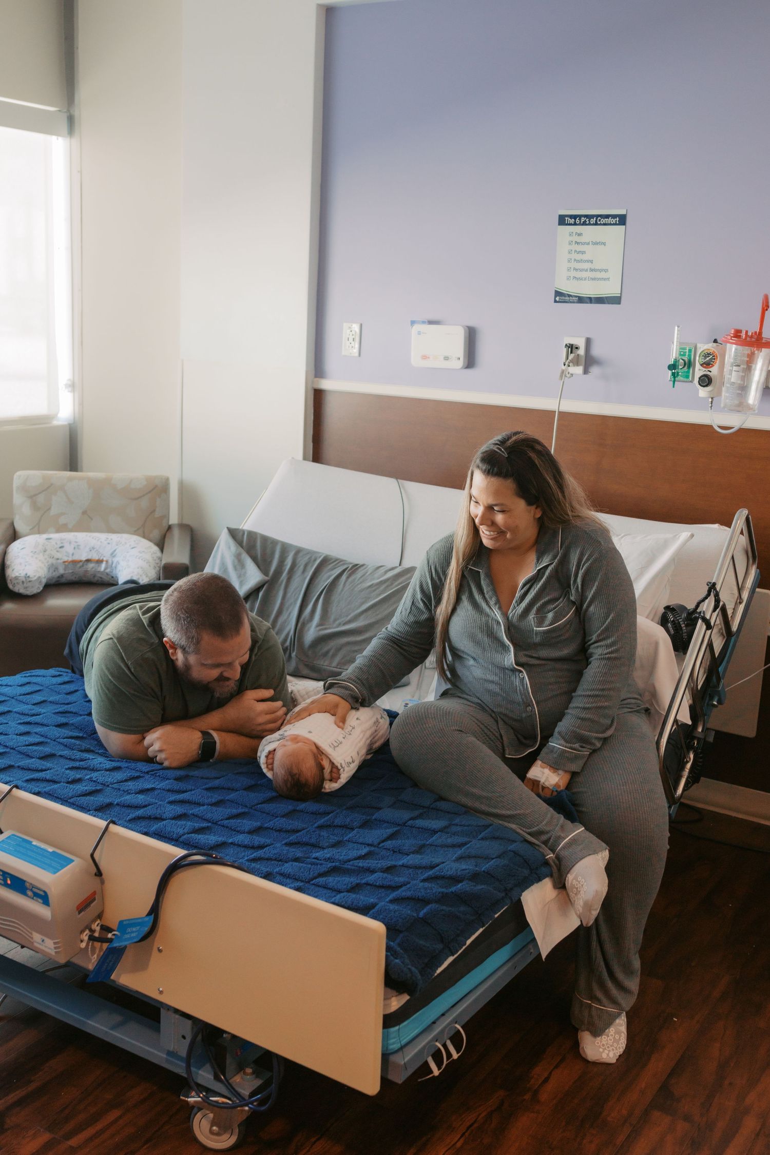 New parents smile at their newborn baby on a hospital bed in a modern maternity ward room.