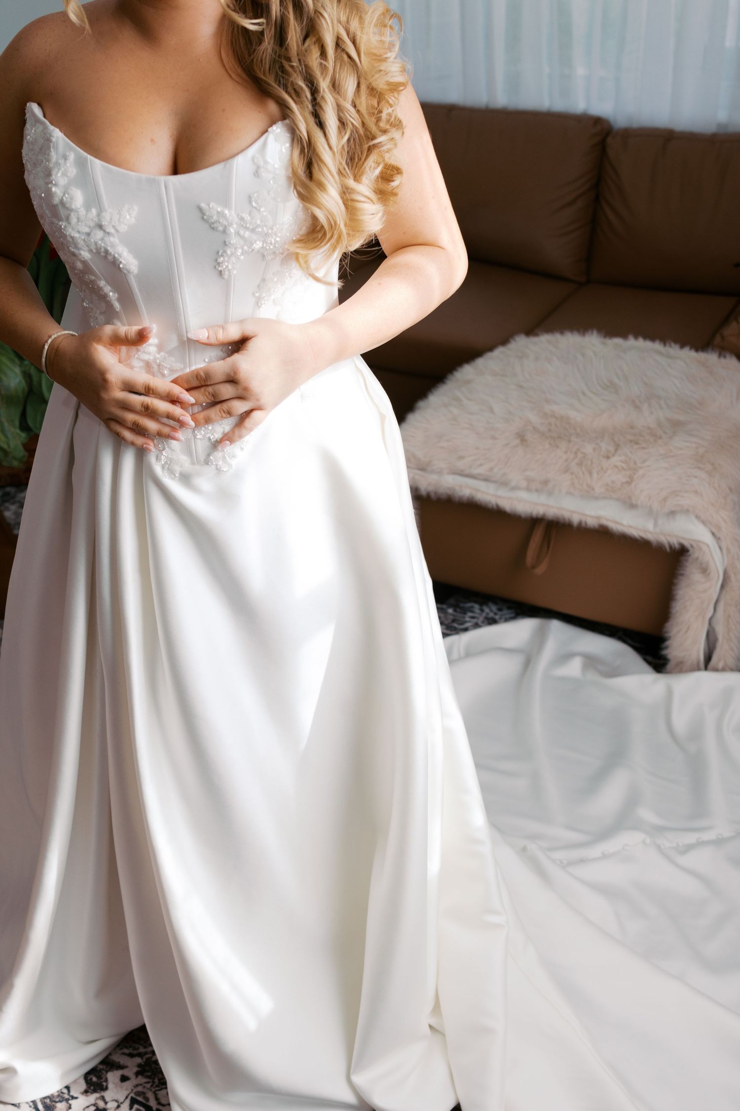 Bride in elegant white satin wedding gown with lace corset bodice, posing indoors near a brown sofa.