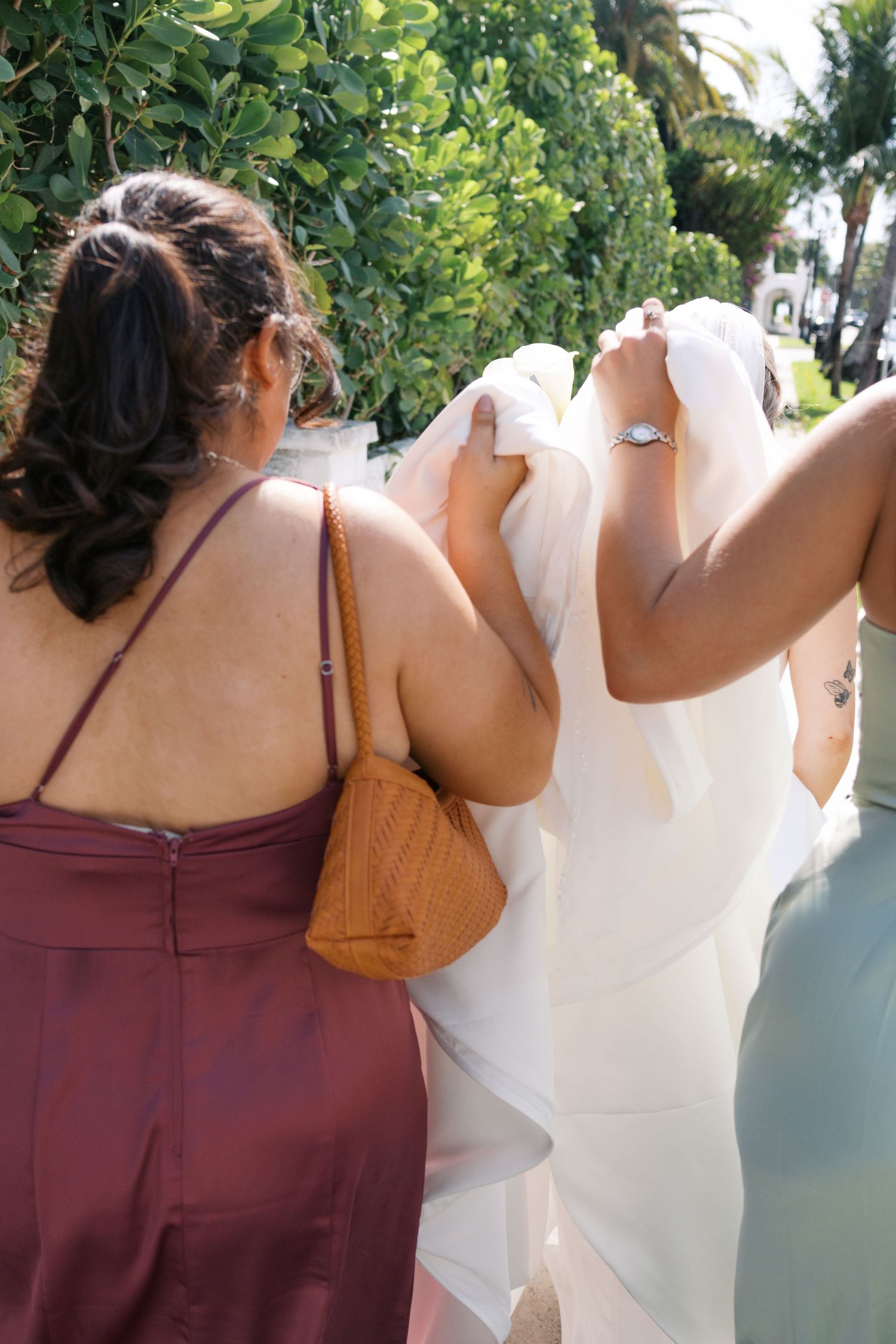 Bridesmaids in burgundy dresses assist bride in white gown and veil walking outdoors on sunny wedding day.