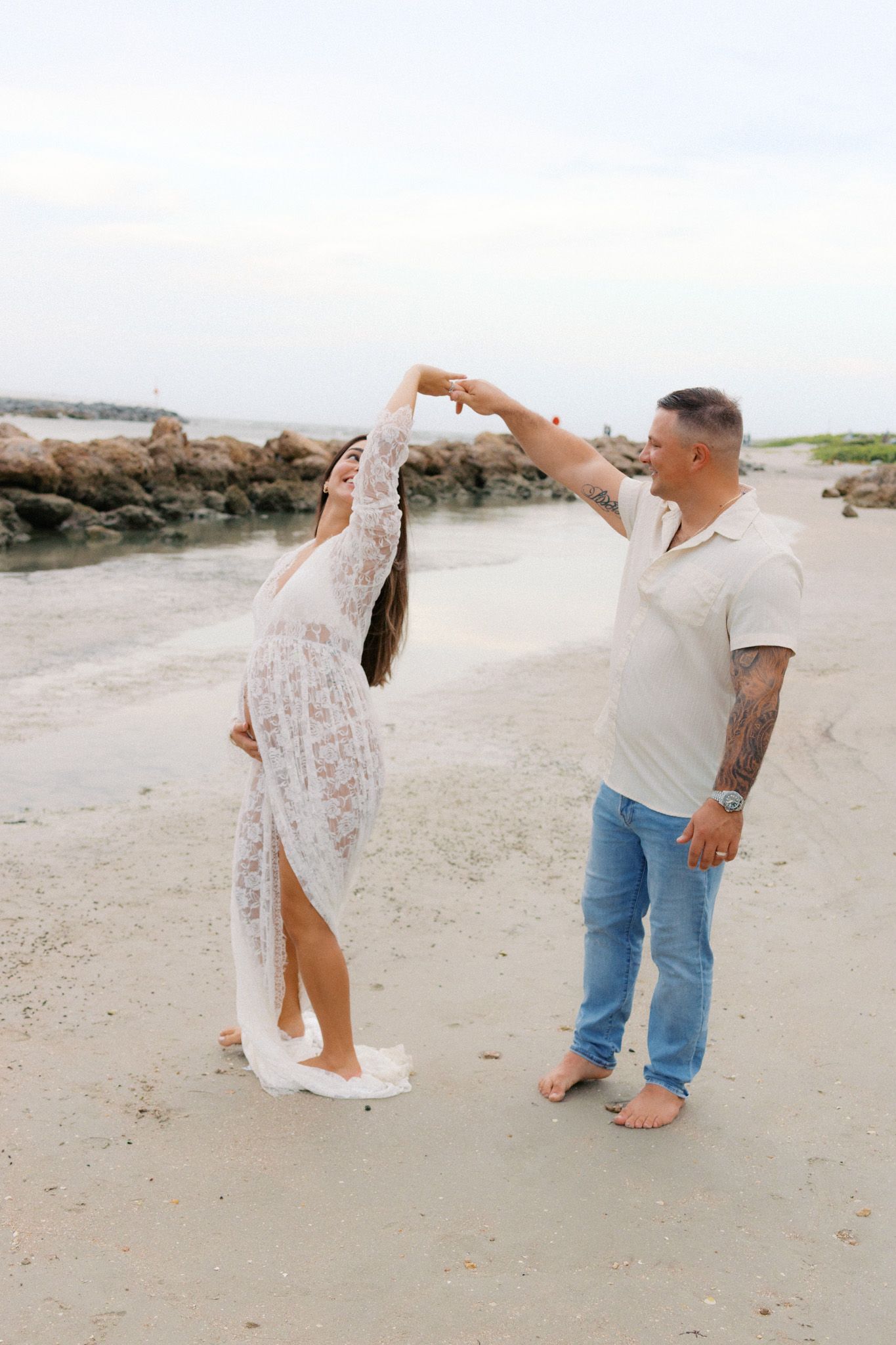 Couple dancing on sandy beach, man spinning woman in white lace dress near rocky shoreline with palm trees.