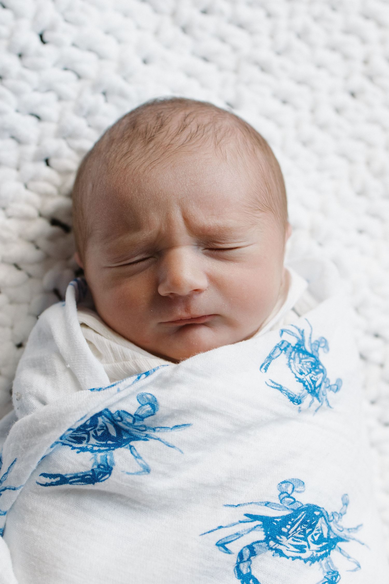 Sleeping newborn baby wrapped in white blanket with blue pattern on textured white background.
