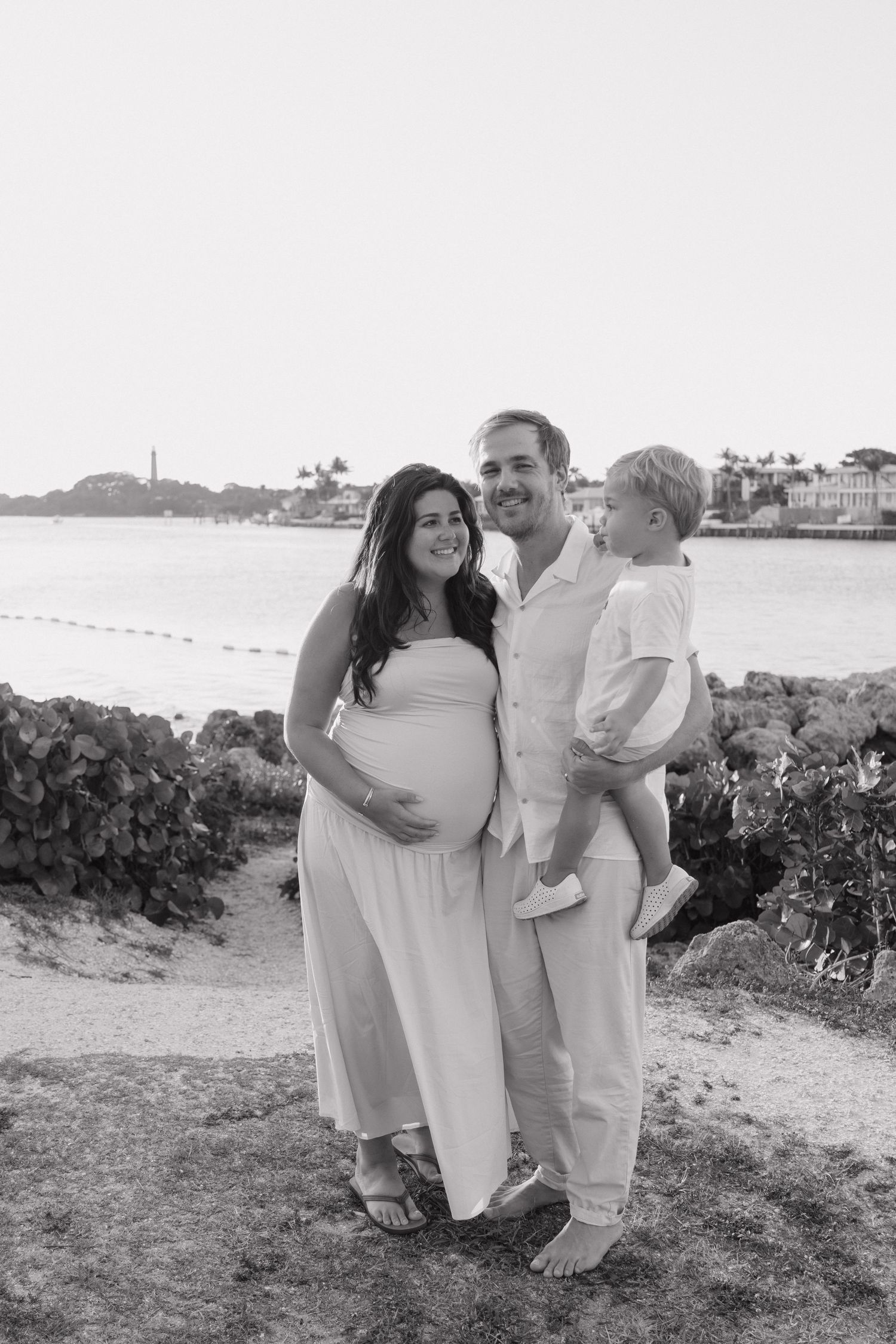 Black and white maternity photo of pregnant couple holding toddler son near waterfront at sunset.