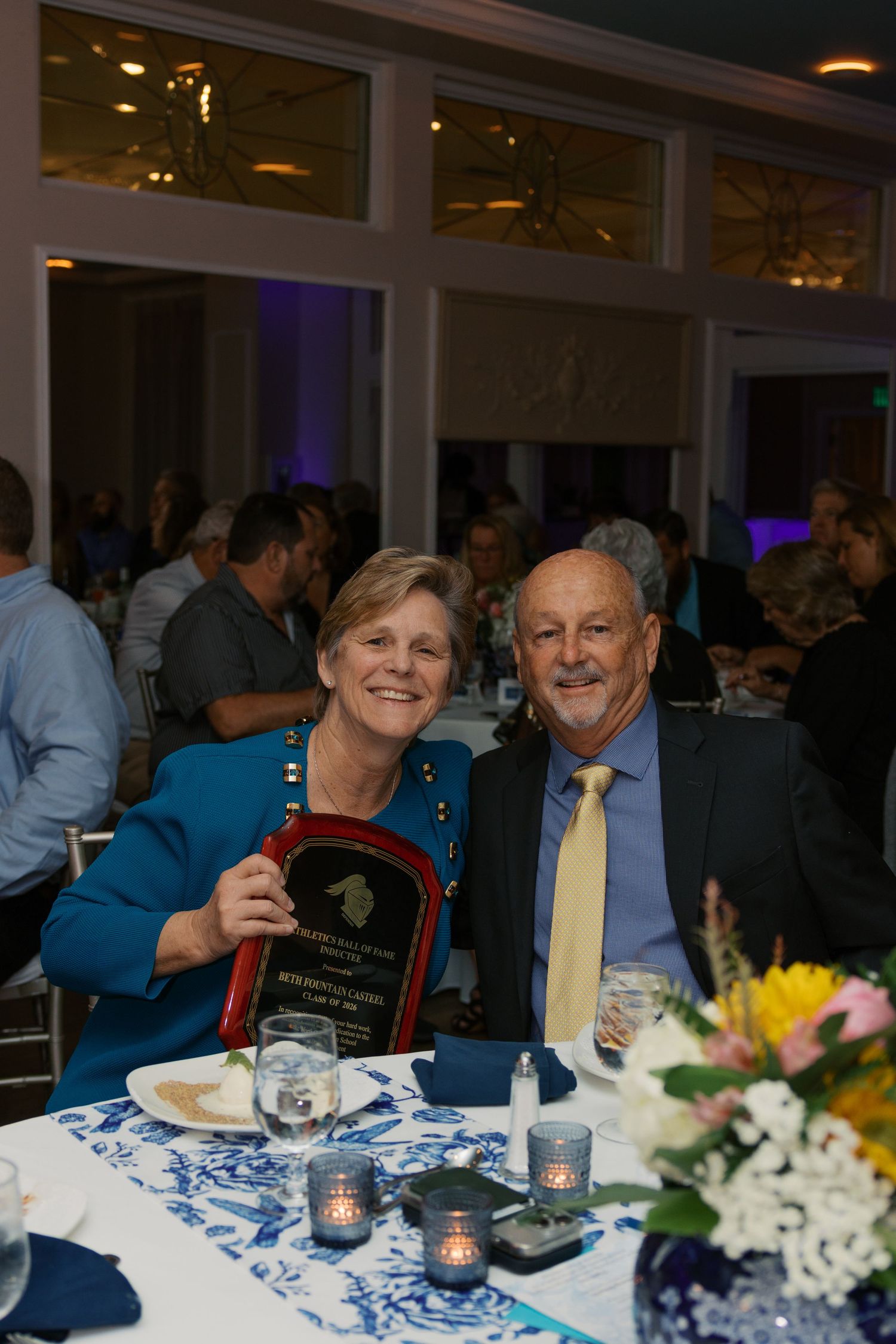Couple smiling at banquet table holding award plaque at formal evening gala event.