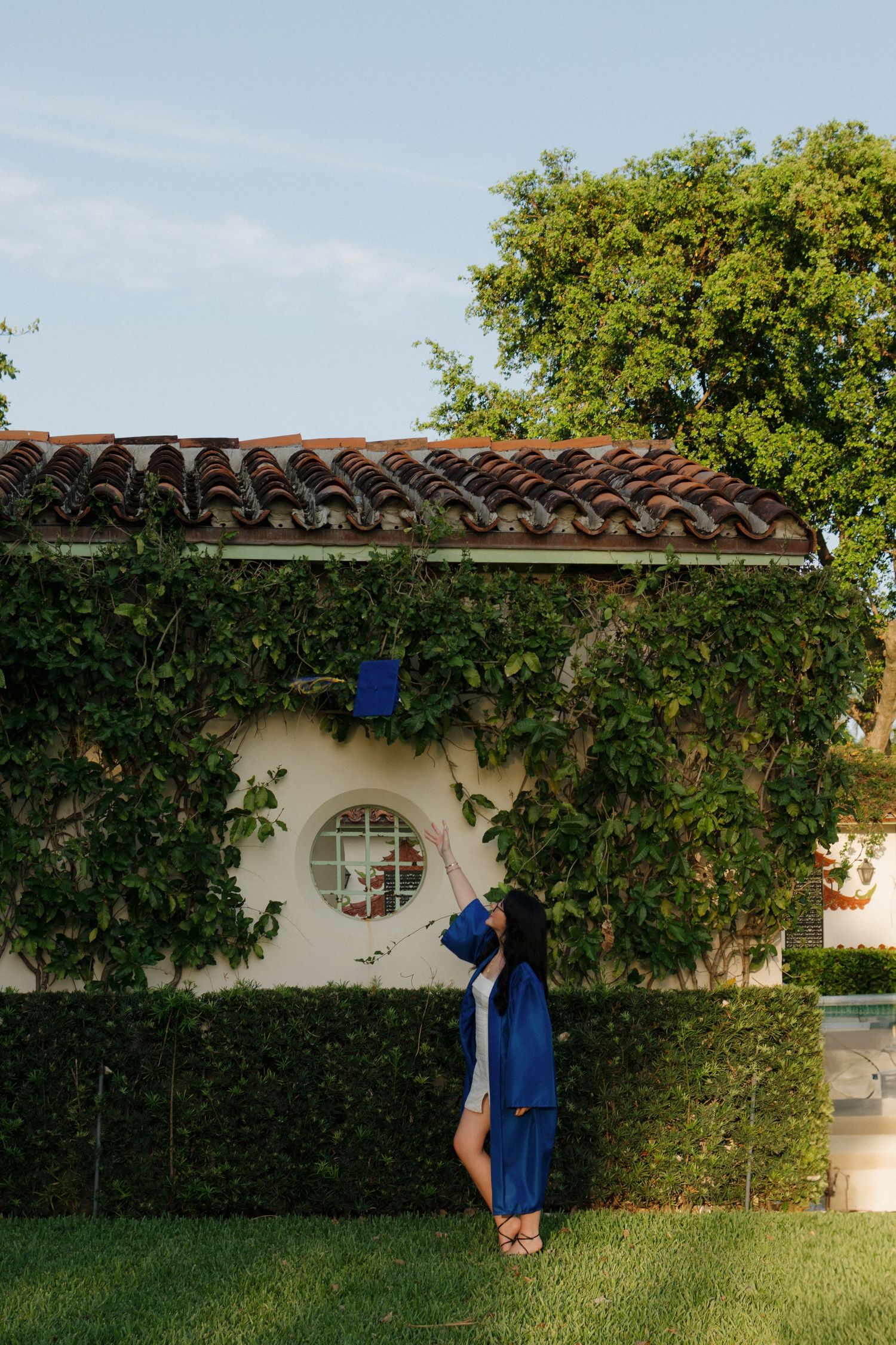 Blue graduation gown contrasts against white wall with circular window and climbing vines.