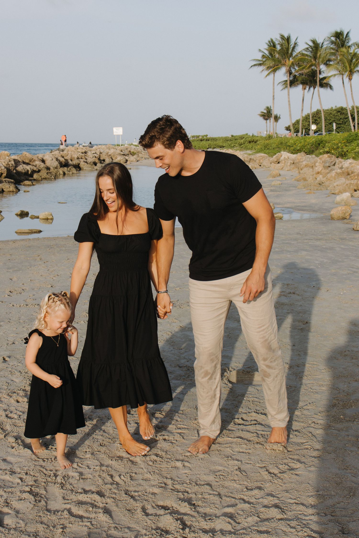 A family walking together on a tropical beach with palm trees and rocks in the background.