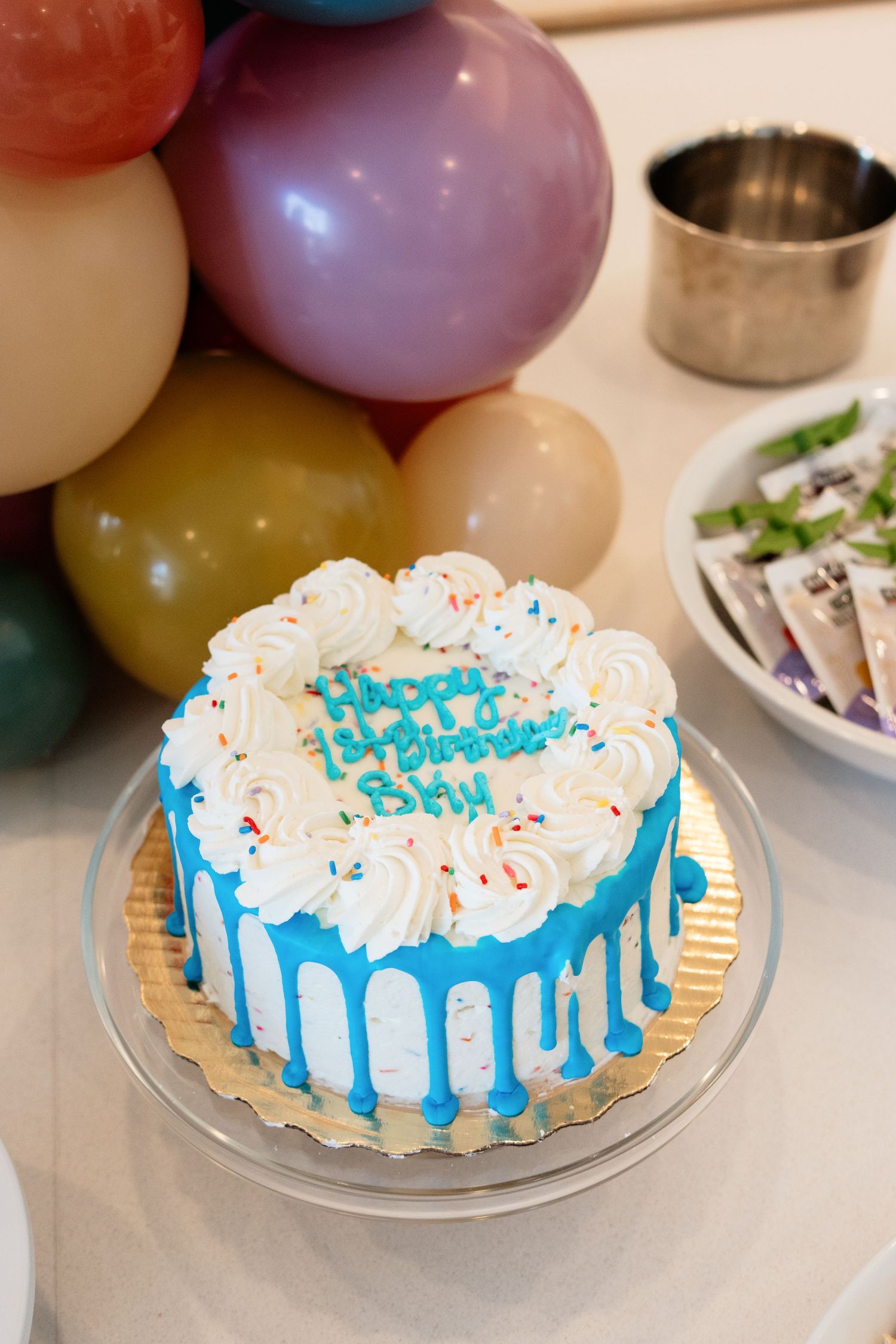 Blue and white Happy Birthday cake with drip frosting and colorful sprinkles on a glass stand with balloons in background.