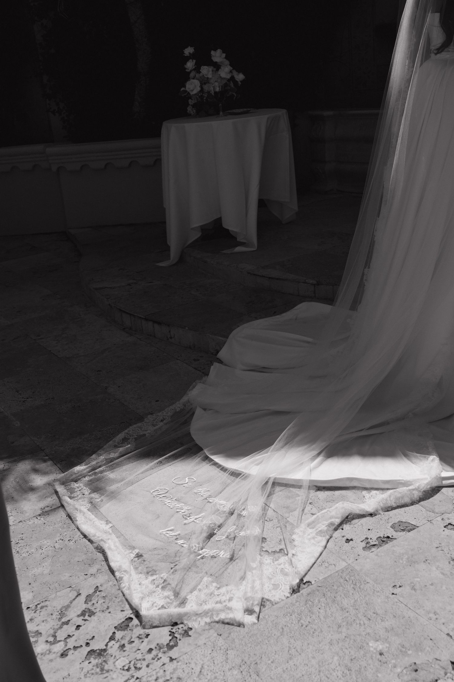 Black and white photo of a wedding dress train and veil spread on stone floor with floral arrangement on table.