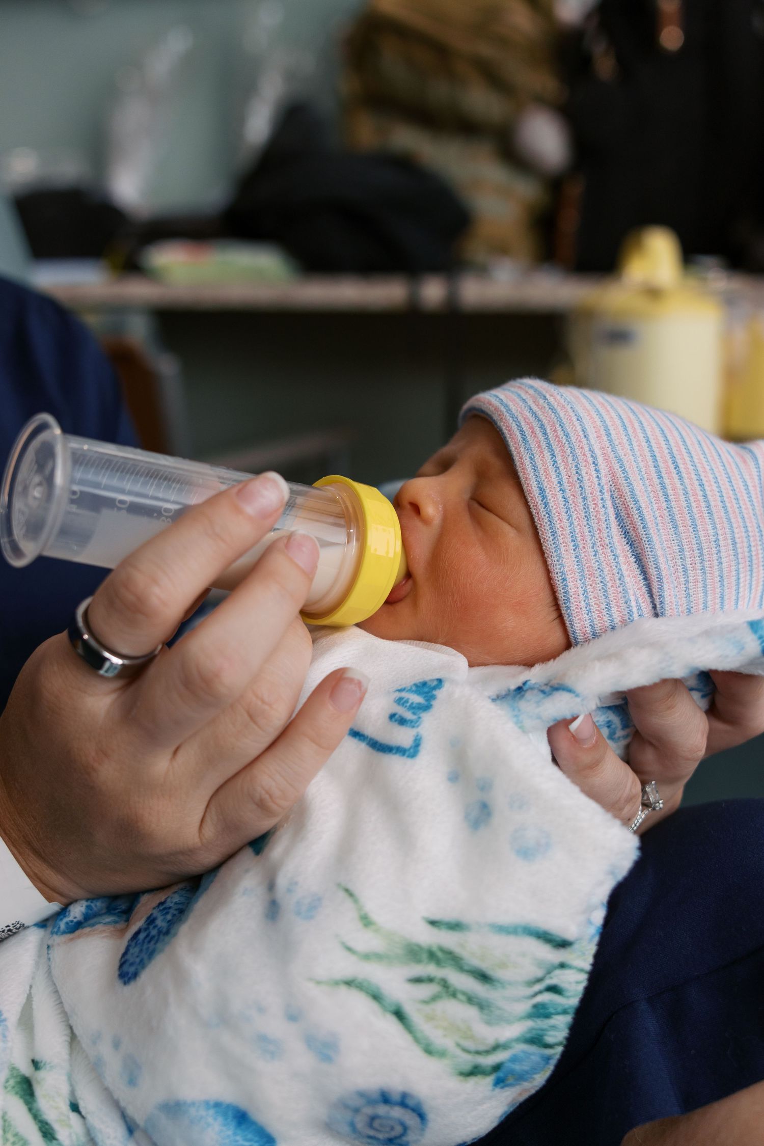 Newborn baby being bottle-fed by mother, wearing striped hat and wrapped in a floral blanket.