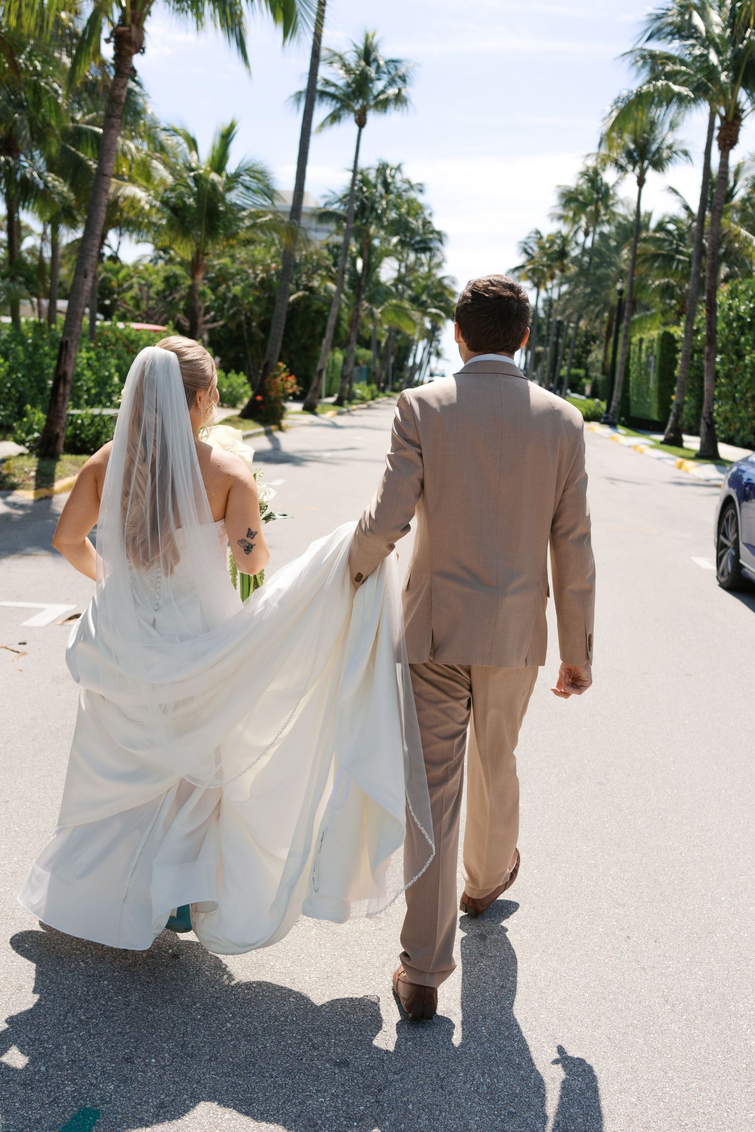Bride and groom walking hand in hand down a palm tree-lined street, bride in white gown with veil, groom in tan suit.