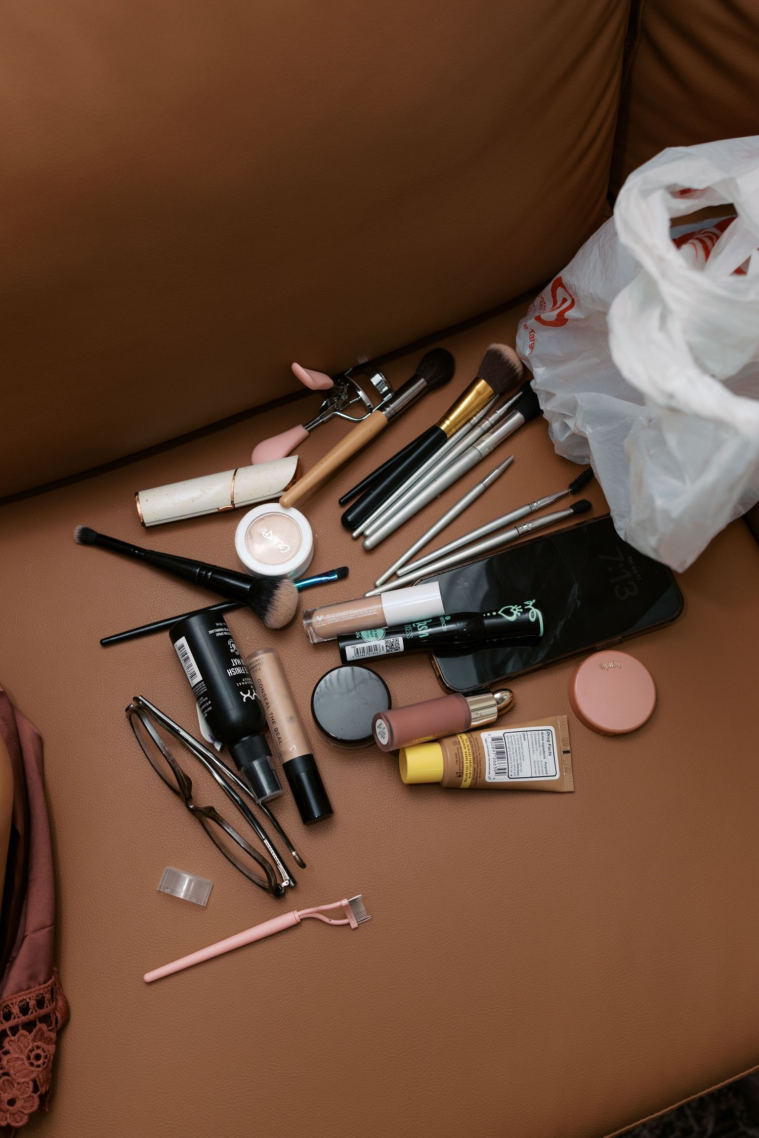 Makeup collection spread on brown leather sofa including brushes, NYX products, foundation, mascara, and eyeglasses.