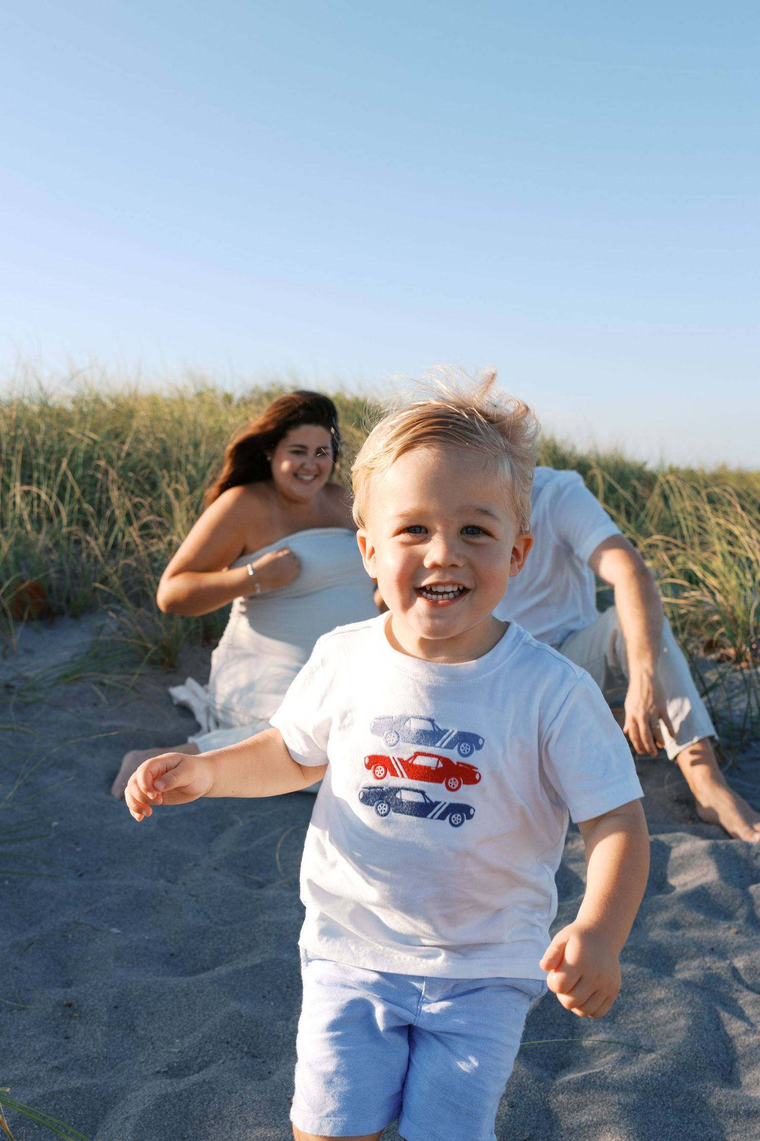 A joyful toddler in a car-print shirt runs on a sandy beach while two women smile and play behind him.