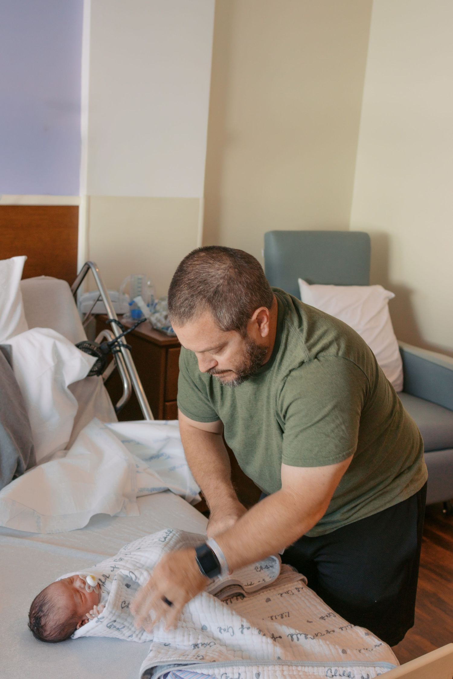 A father tenderly swaddles his newborn baby on a hospital bed in a maternity ward room.