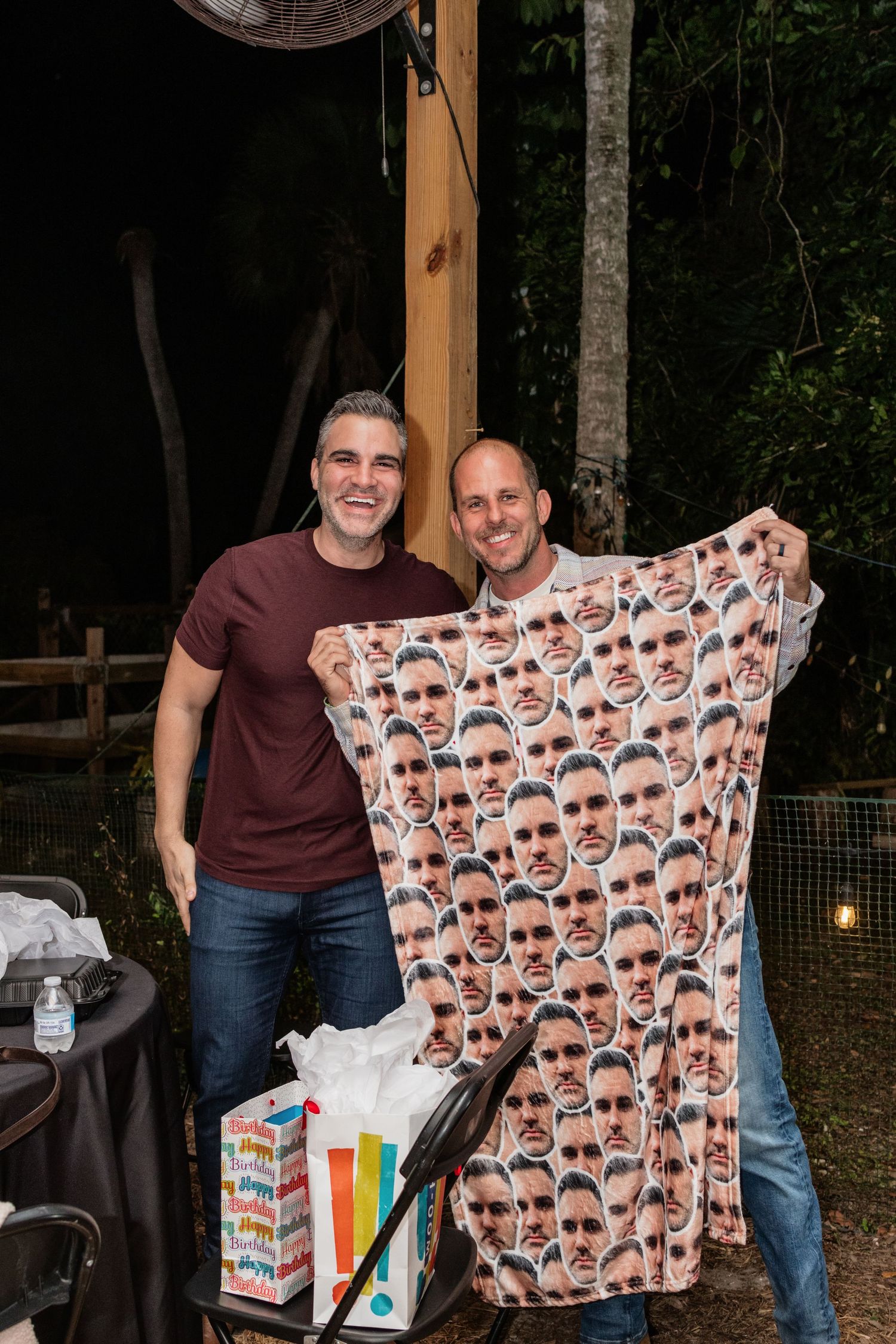 Two men smiling and holding a custom blanket covered in repeated face prints at an outdoor party.