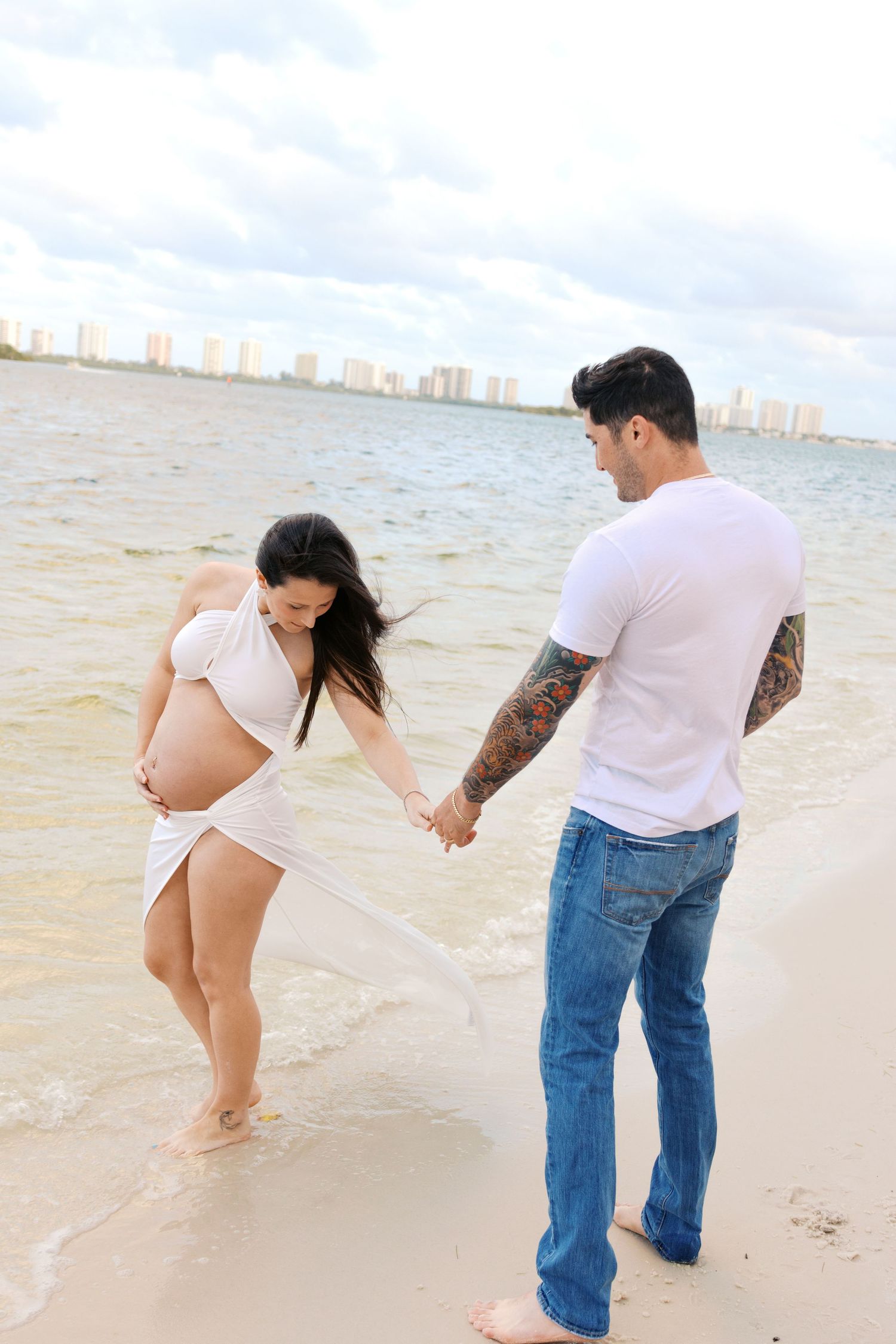 Pregnant couple holding hands on a beach, woman in white wrap dress cradling baby bump, man in white tee and jeans with tattoos.