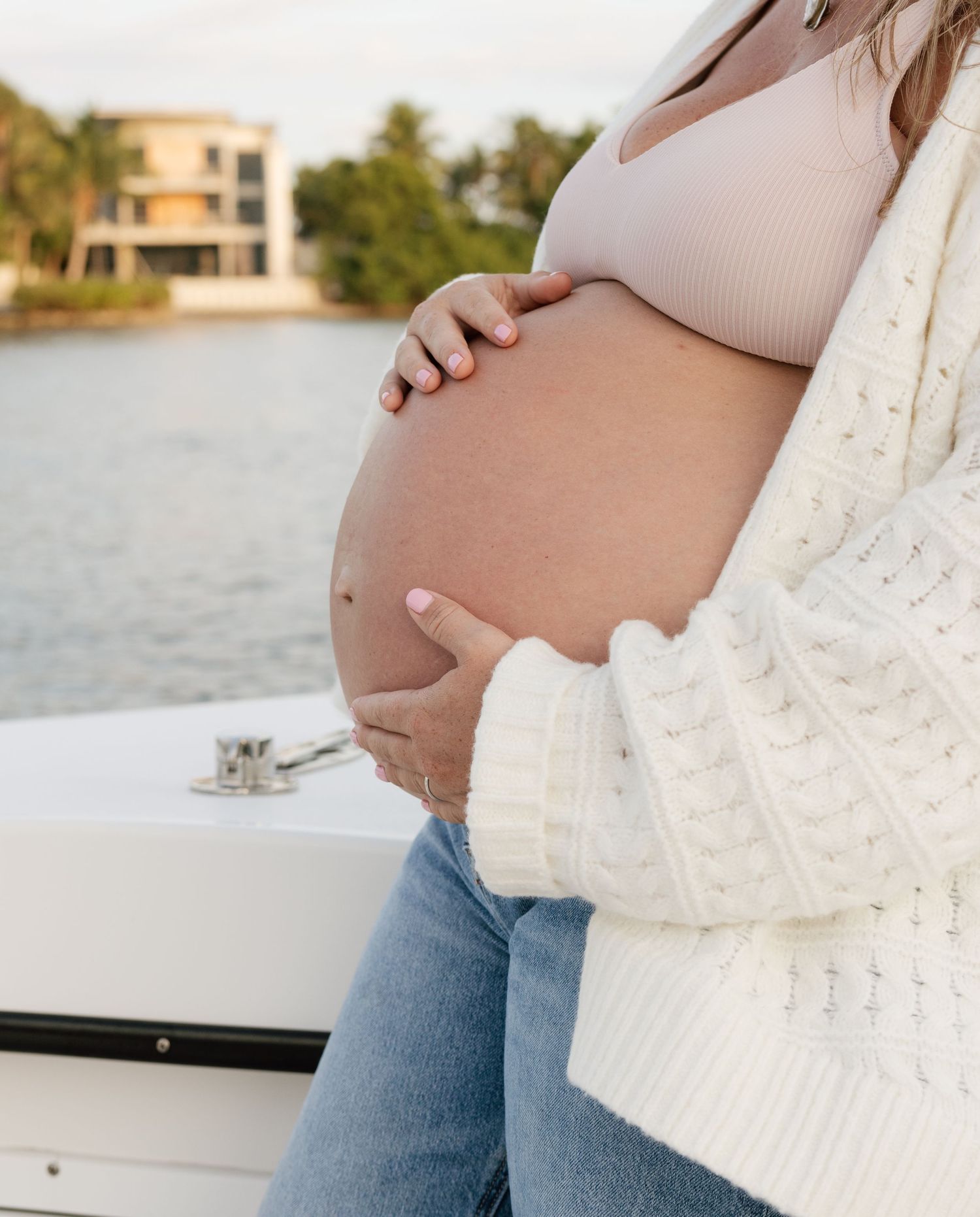 A pregnant woman in a white sweater and jeans sits on a boat deck overlooking the water.
