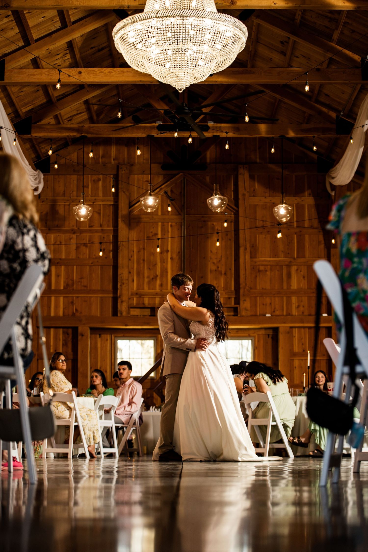 Bride and groom share first dance under crystal chandelier in elegant rustic barn wedding reception.