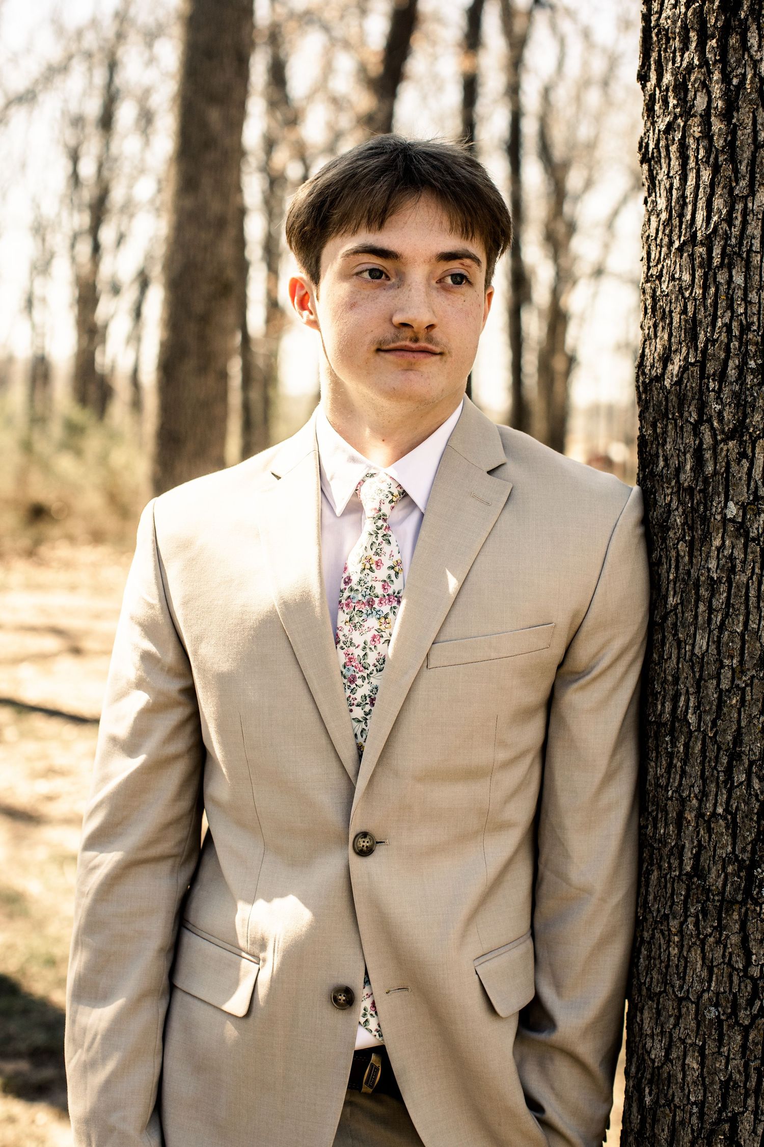 Young groom in tan suit with floral tie leaning against tree in outdoor woodland setting.
