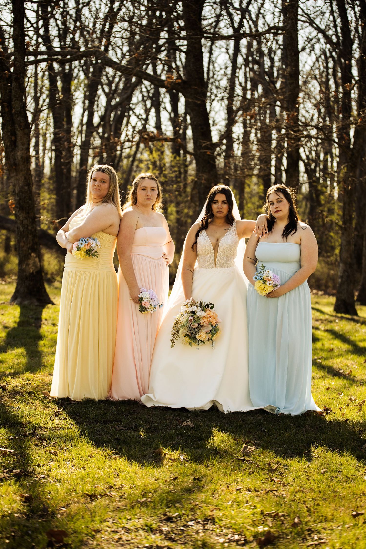 Bride poses with three bridesmaids in pastel gowns holding bouquets in a sunny wooded outdoor setting.