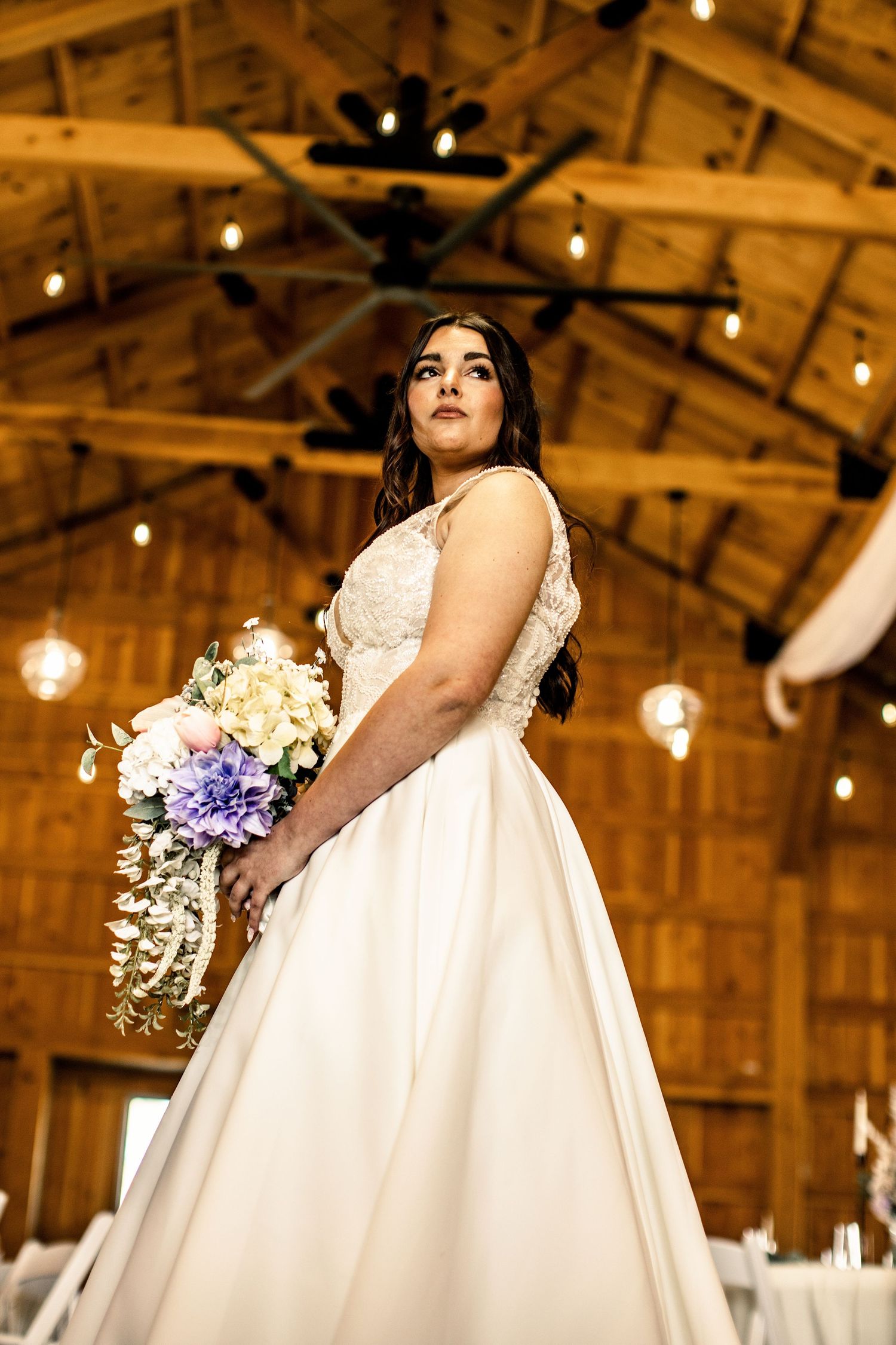 Bride in white ball gown holding colorful bouquet inside a rustic barn wedding venue.