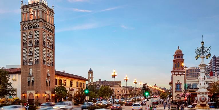 Historic brick clock tower and buildings line a bustling city street under a bright blue sky.