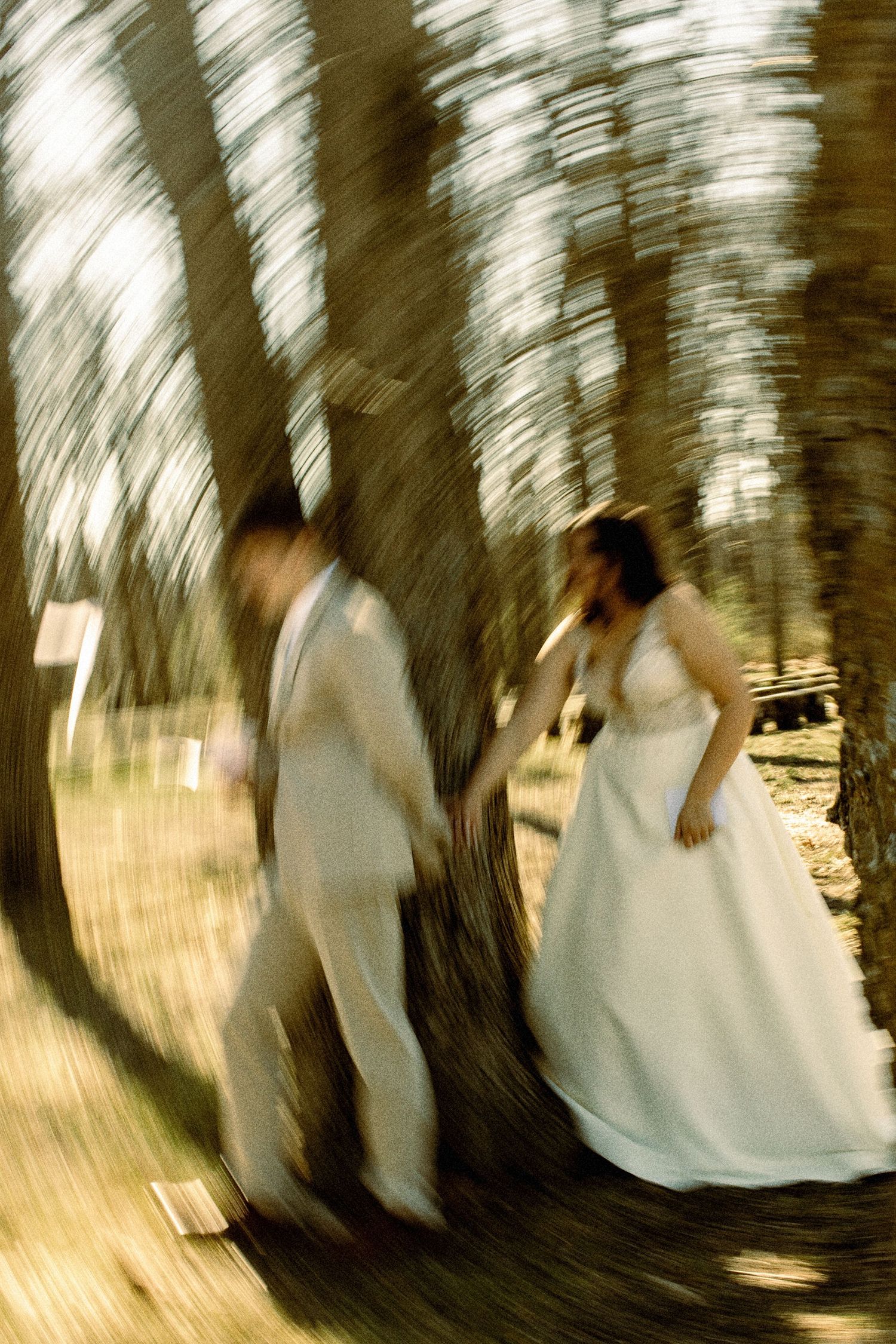Artistic blurred motion photo of bride and groom walking among tall trees outdoors.