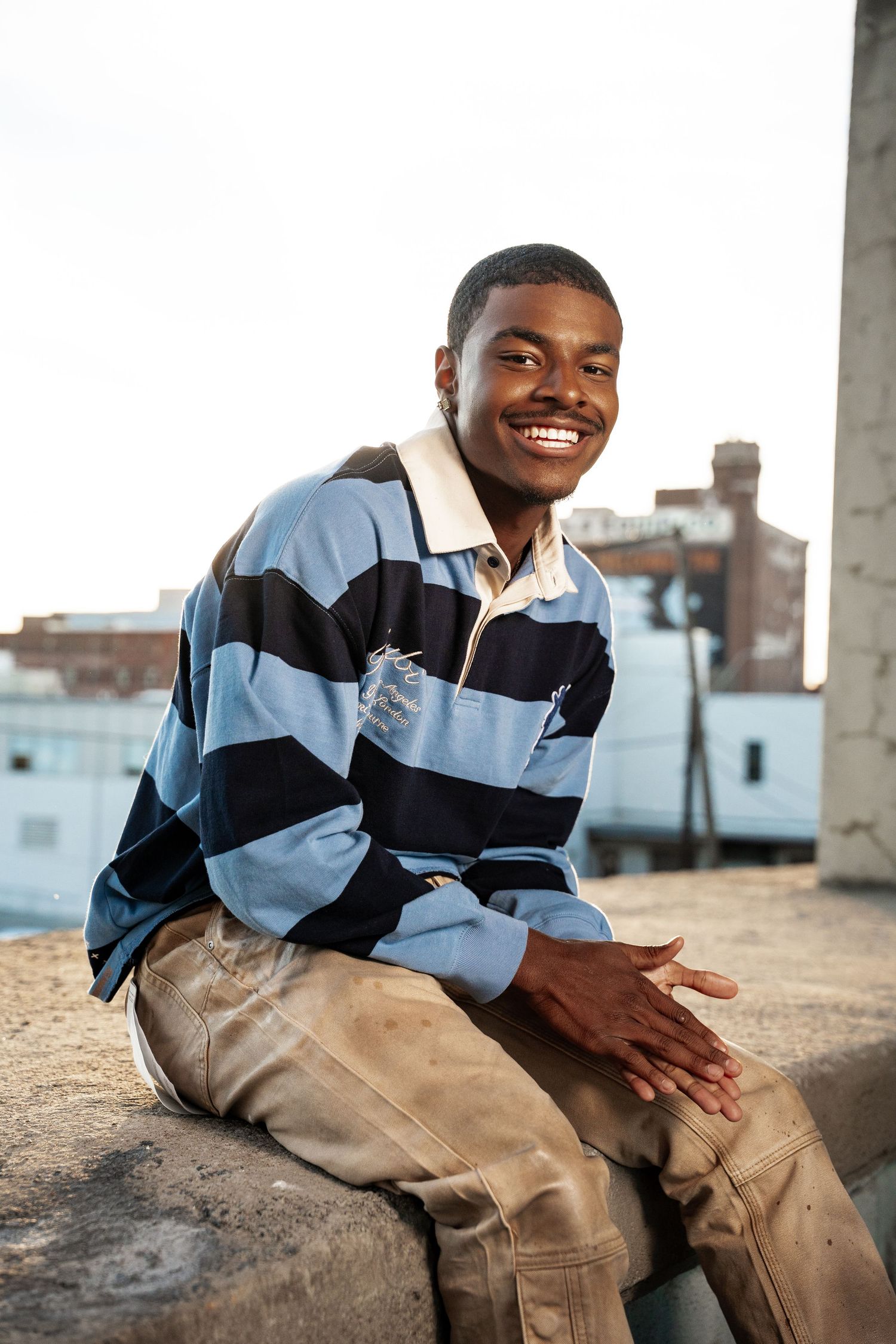 Person in striped polo and khaki pants sitting outdoors with joyful expression.