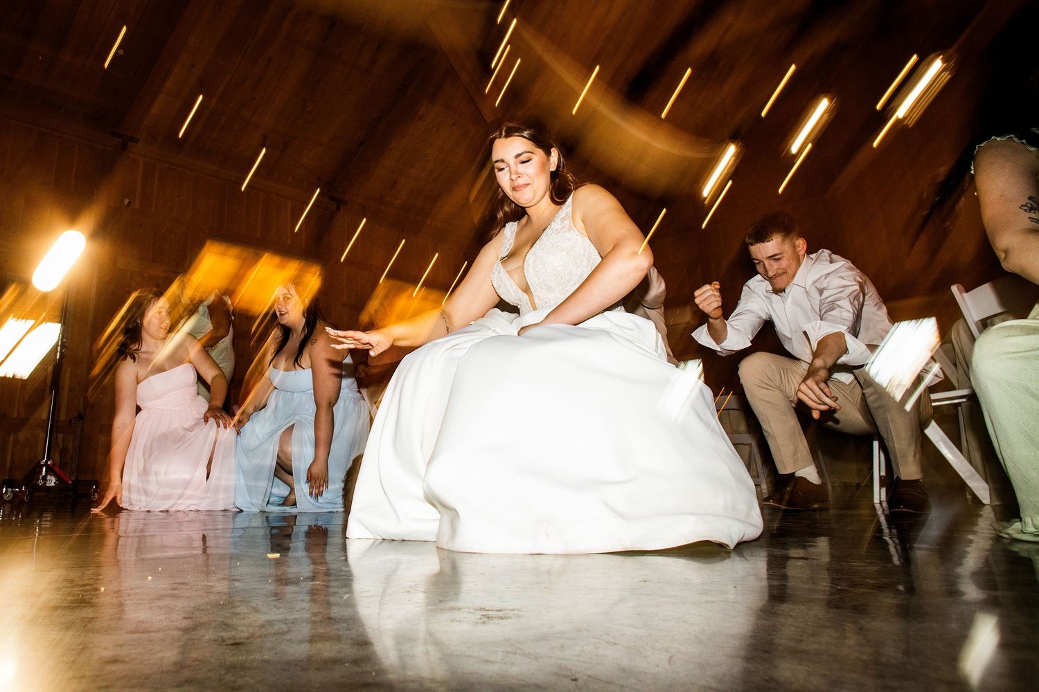 Bride in white gown dancing joyfully with guests during lively wedding reception in a warmly lit barn.