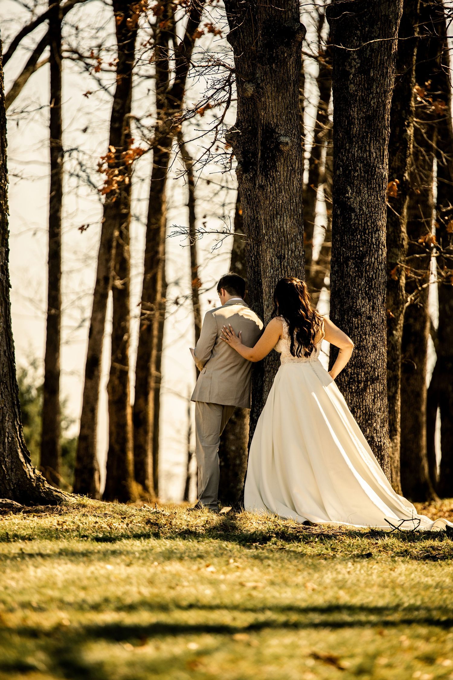 Bride and groom standing together among tall autumn trees in golden evening light.