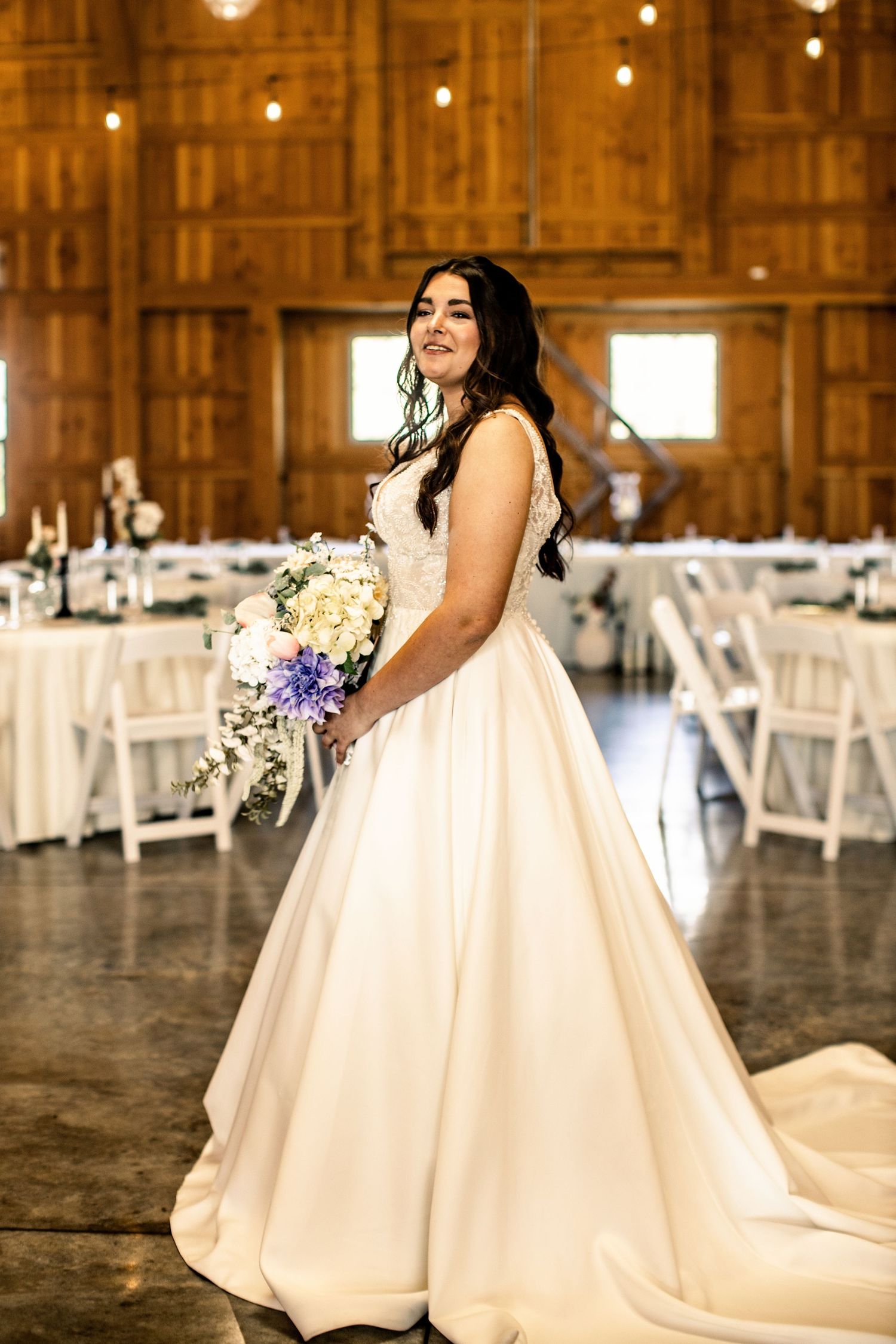 Bride in ball gown wedding dress holding purple and white bouquet inside rustic barn reception venue.
