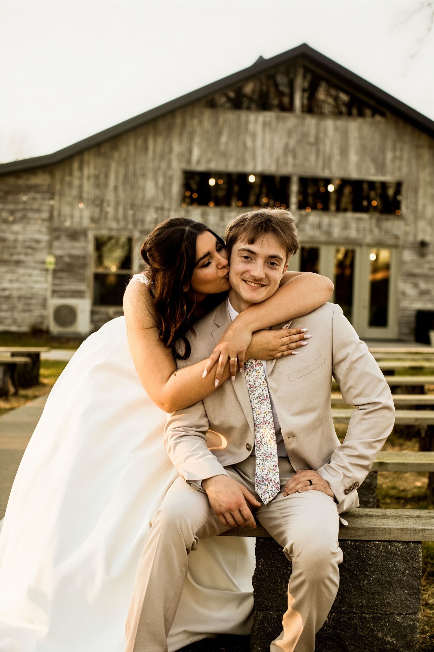 Bride kissing groom on cheek while embracing outside rustic barn wedding venue on their wedding day.