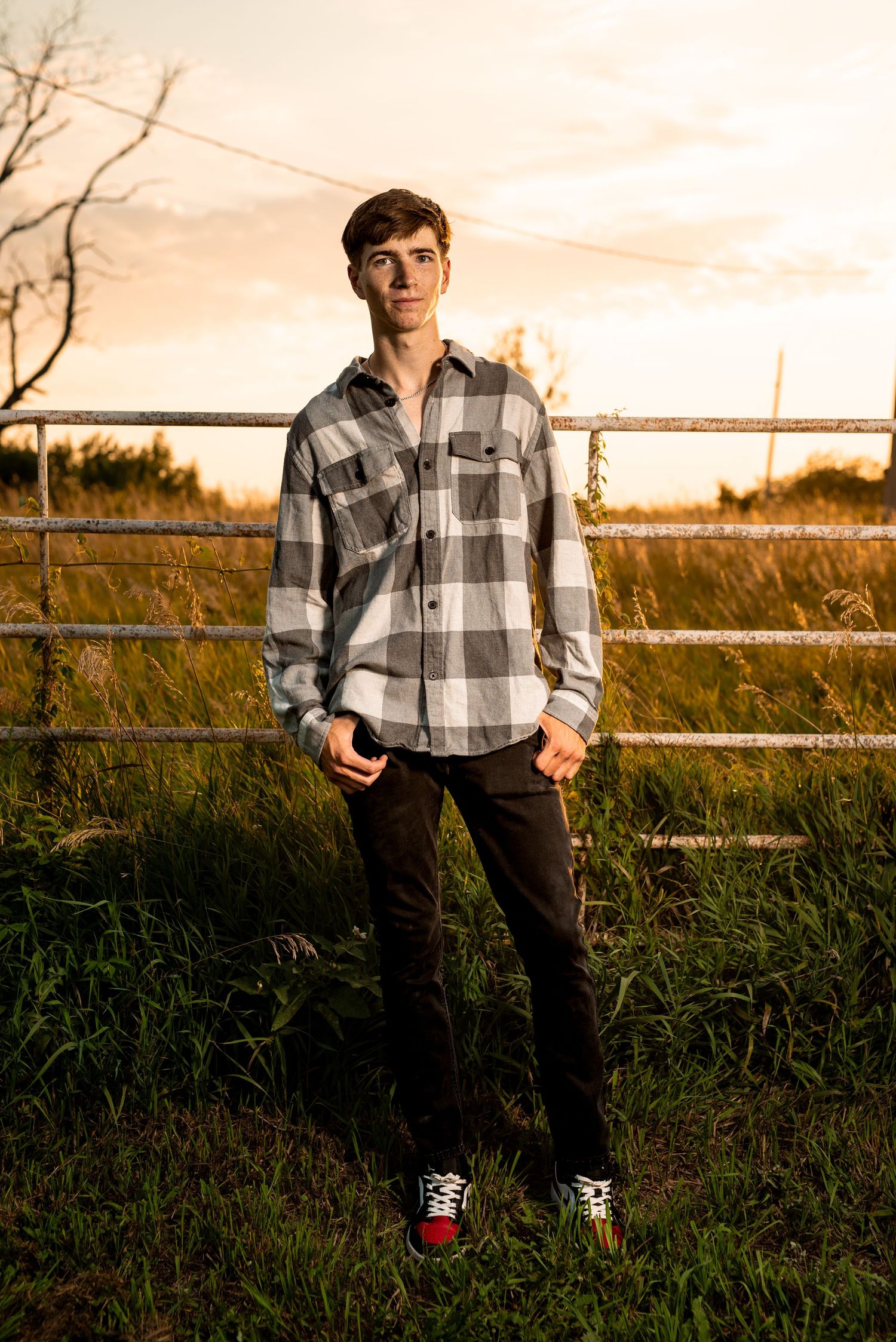 Senior portrait in plaid flannel shirt against rustic fence during golden hour.
