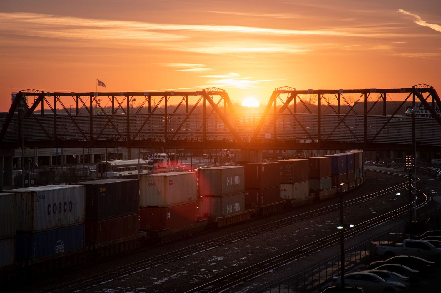 A dramatic orange sunset silhouettes a railway bridge and cargo containers in an industrial rail yard.