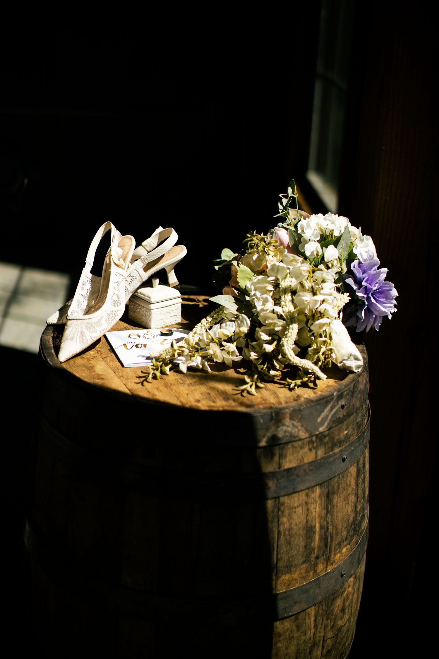 Wedding shoes, bouquet of white and purple flowers, and rings displayed on a rustic wooden barrel in soft natural light.