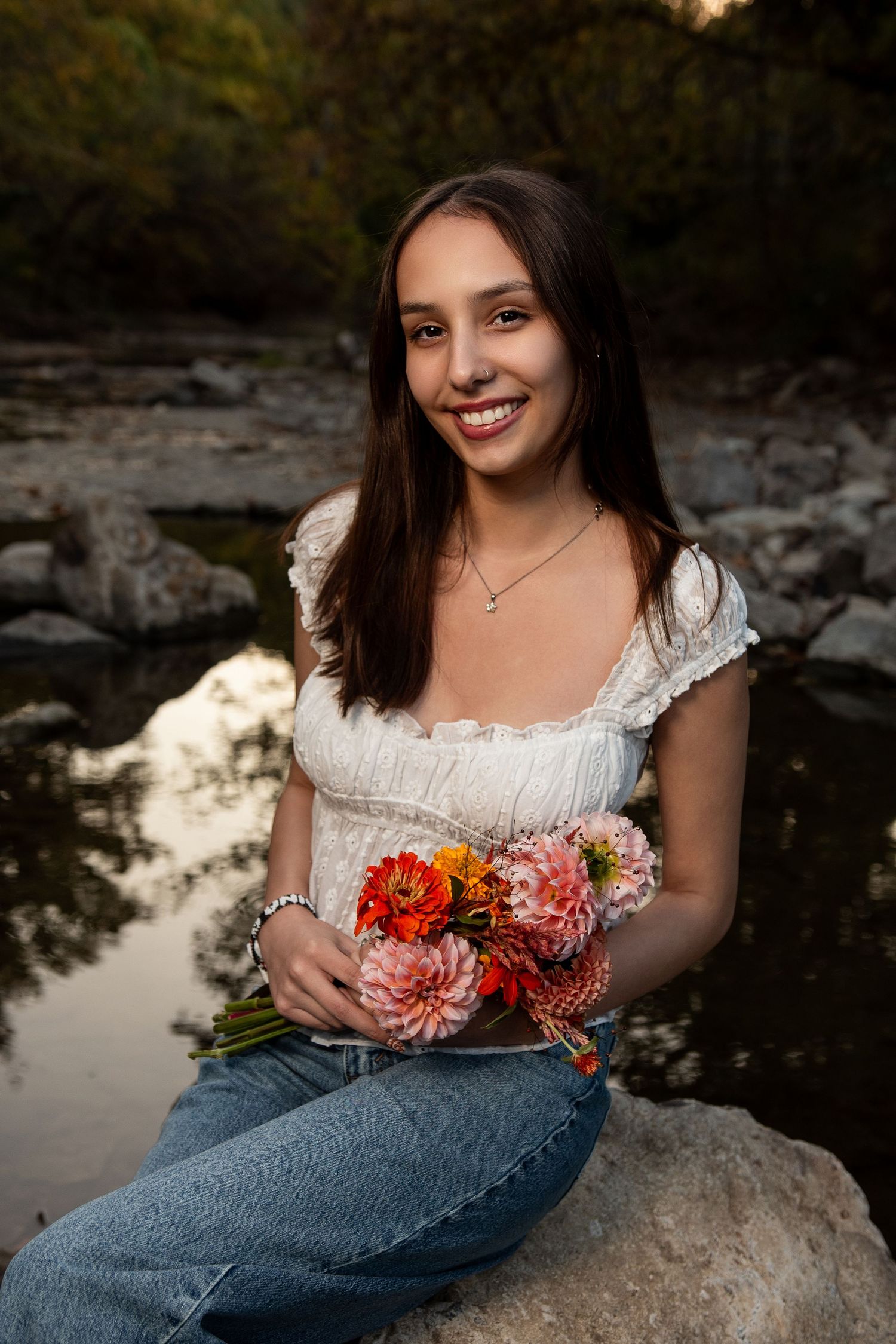 Person in white top holding vibrant flowers while sitting on rocks near water at sunset.