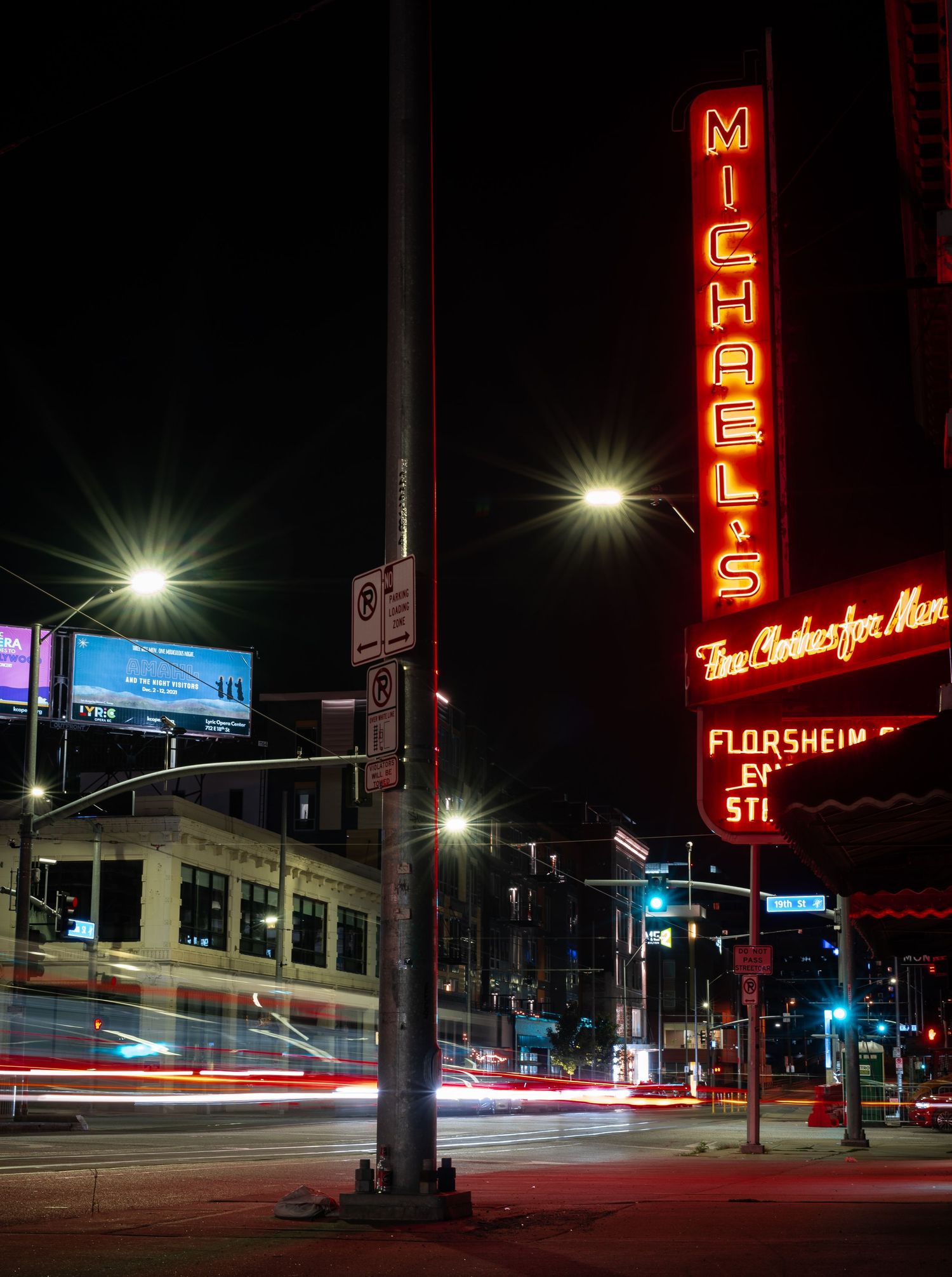 Long exposure night photography captures red neon signs and light trails along a dark urban street corner.