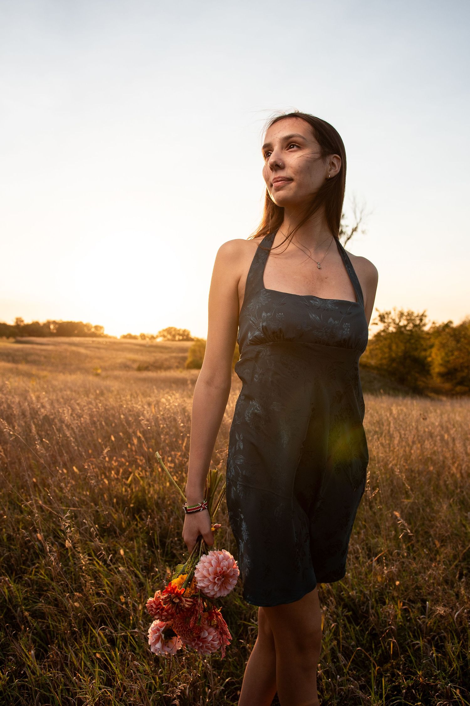 A woman in a black halter dress holds fresh flowers while standing in a golden field at sunset.
