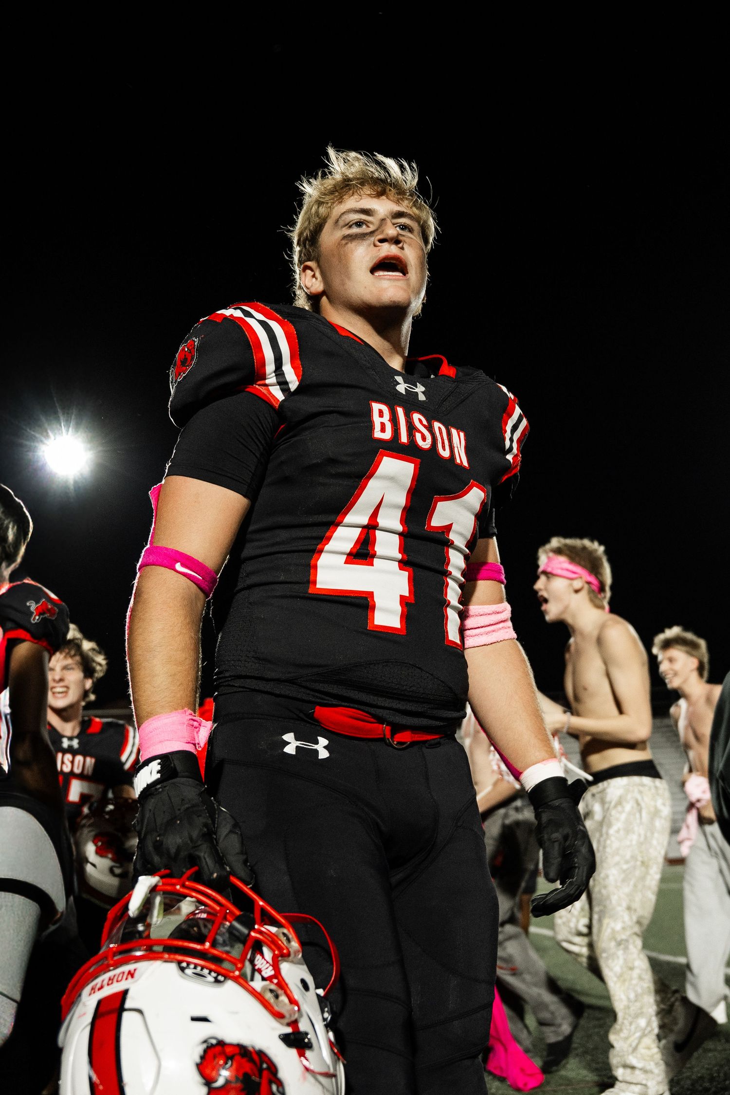 Football players in black Bison uniforms with pink accents standing on field at night.