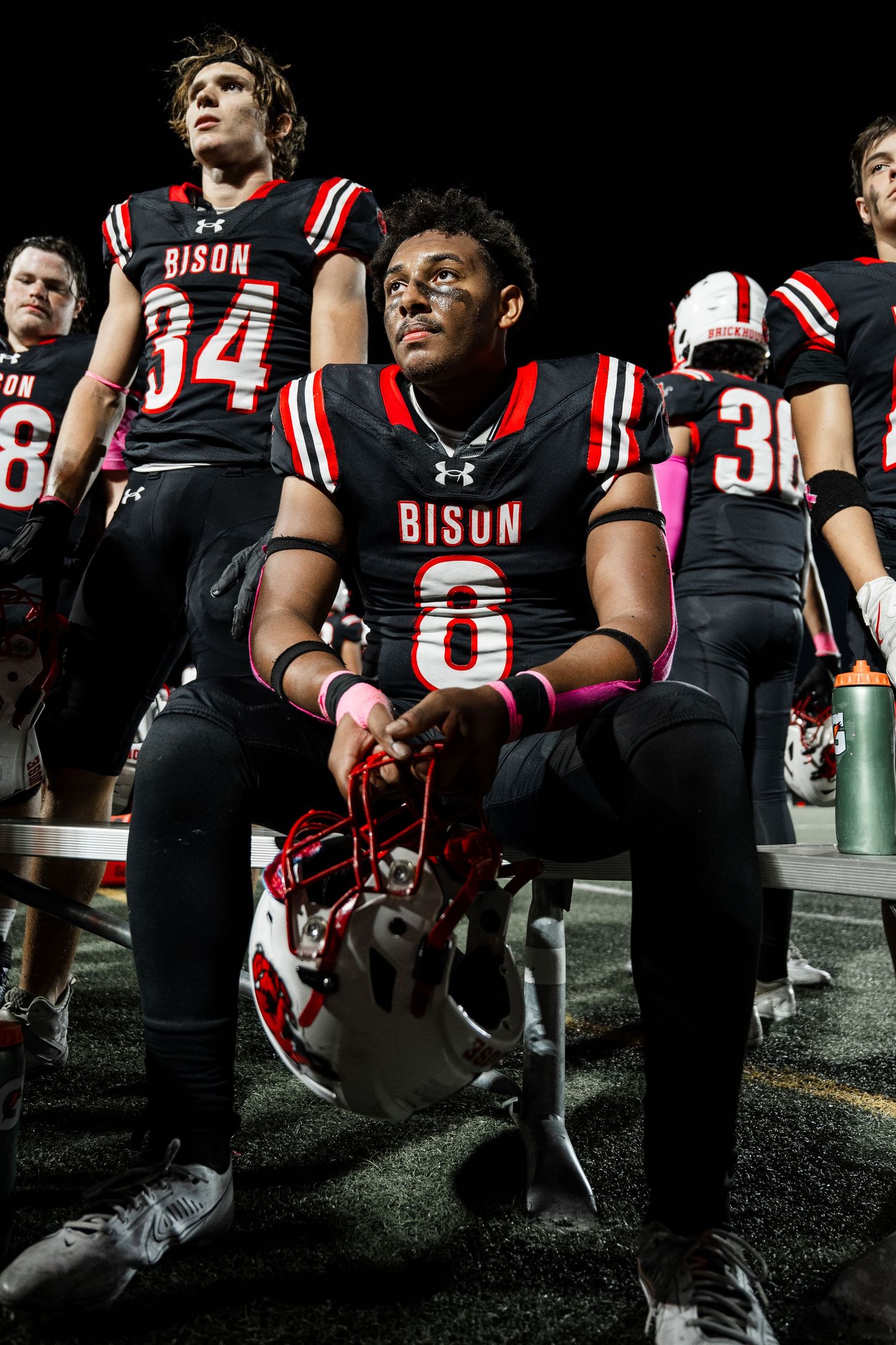 Football team in black and red uniforms sitting on sideline bench during night game.