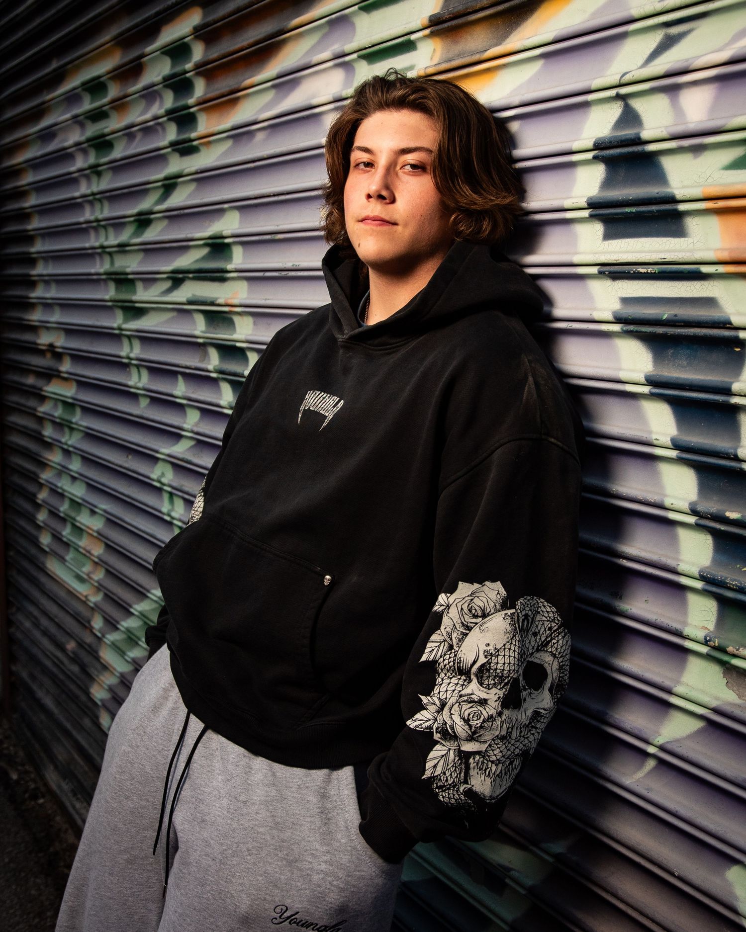 Person in black hoodie poses against industrial metal backdrop with dramatic lighting.